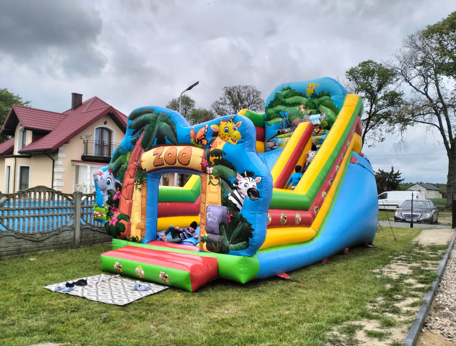 Kolorowy dmuchany zamek ze zjeżdżalnią w motywie zoo, dzieci bawią się w środku. Na trawie leżą buty, w tle dom i samochód. Pochmurne niebo.