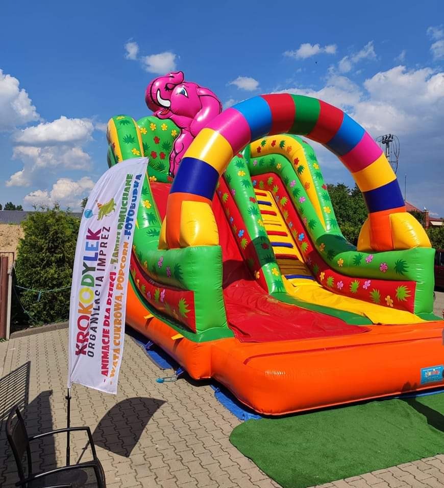 Kolorowa, dmuchana zjeżdżalnia z motywem słonia na zewnątrz, obok flaga reklamowa z nazwą firmy i ofertą: animacje, dmuchańce, wata cukrowa, popcorn, fotobudka.