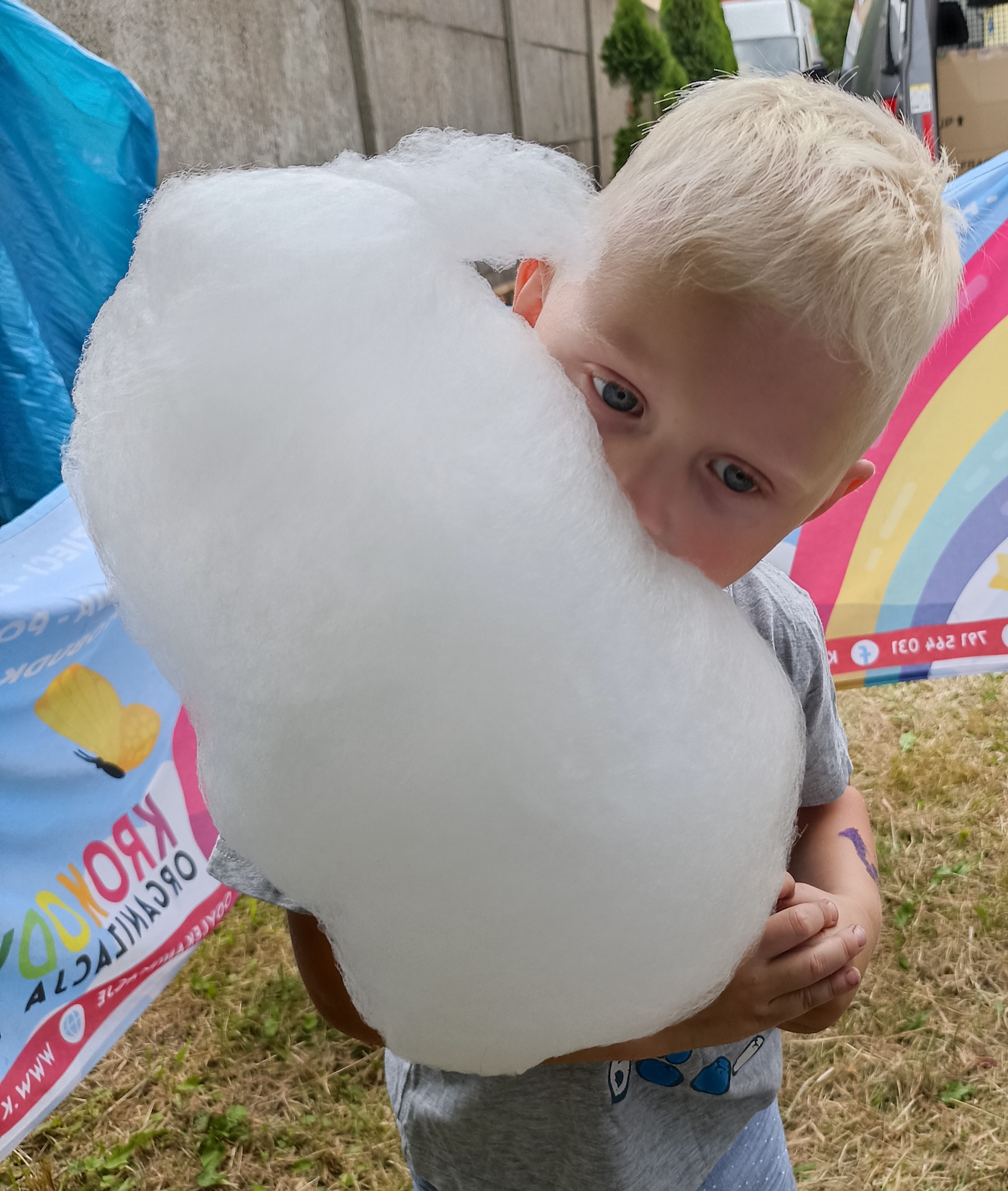 Blondwłosy chłopiec z bliska, trzymający ogromną watę cukrową na tle kolorowego baneru i trawy. Widoczne niebieskie oczy dziecka.