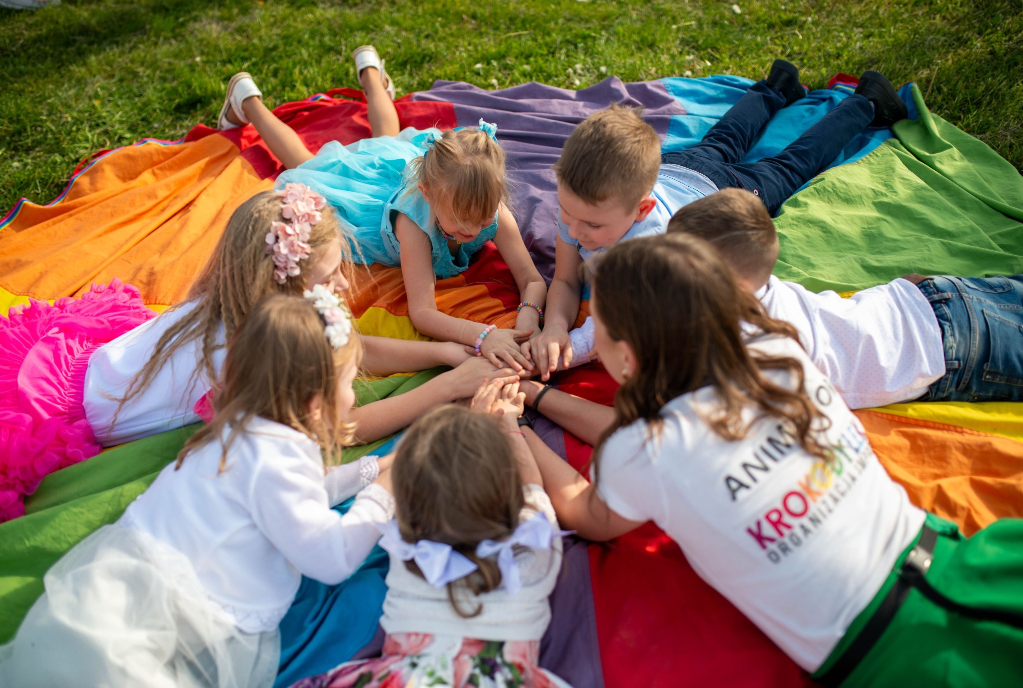 Grupa dzieci w strojach leży na tęczowym kocu, trzymając się za ręce. Widoczna koszulka z napisem 'Animoksy'.