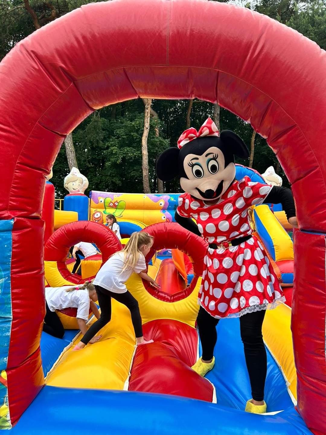 Dzieci bawią się w dmuchanym zamku z animatorem przebranym za postać z bajki. Kolorowy zamek z tunelem i zjeżdżalnią, w tle drzewa.