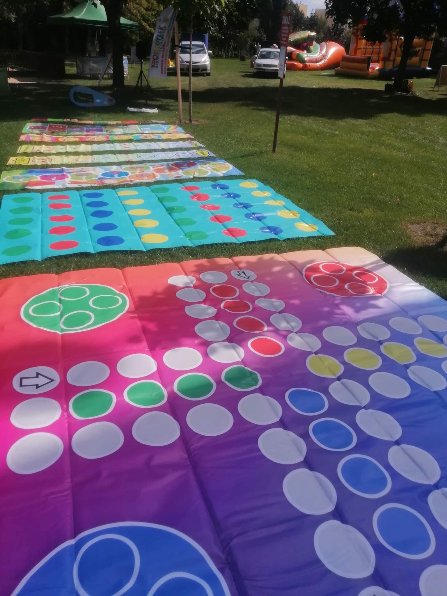 Rozłożone na trawie kolorowe maty do gier zespołowych, w tle dmuchany zamek i inne atrakcje plenerowe. Widok z góry, słoneczny dzień.