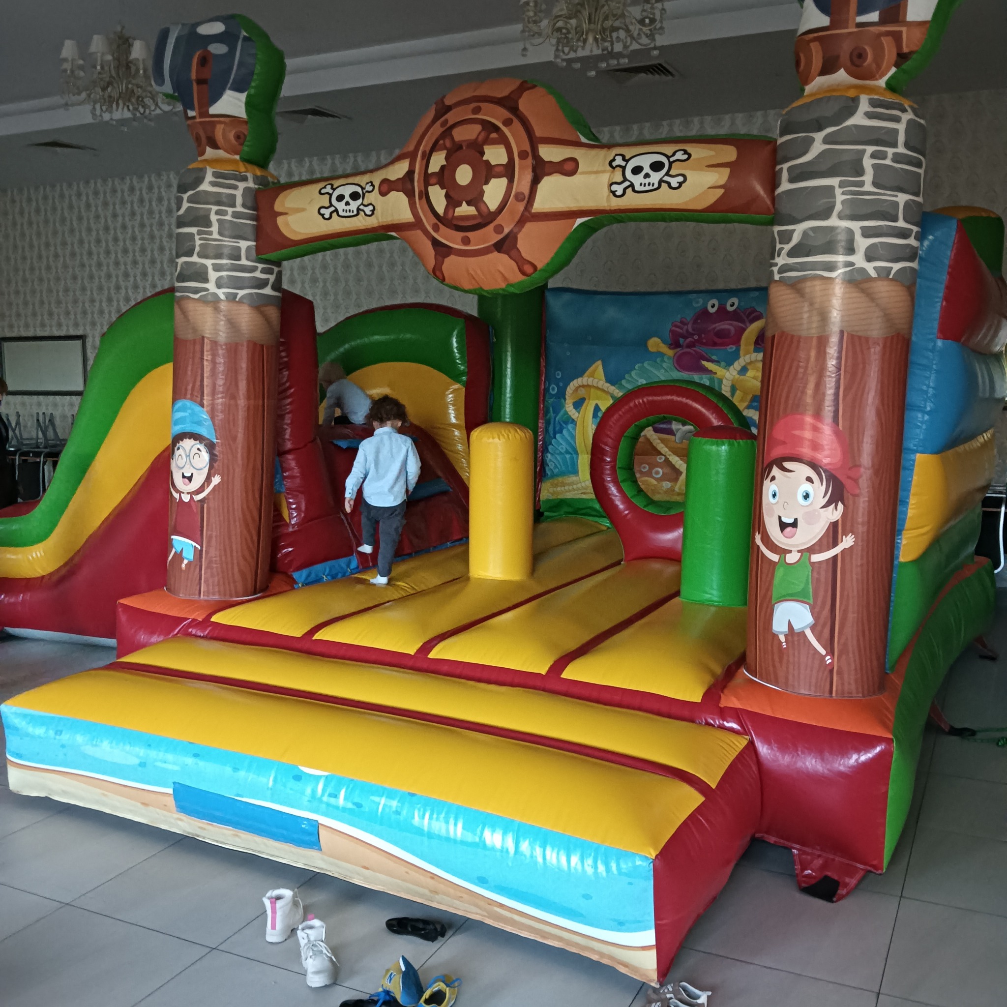 Kolorowy dmuchany zamek w stylu pirackim z motywem czaszek i steru, dzieci w środku. Widoczne buty dziecięce na podłodze. Sala z żyrandolem w tle.
