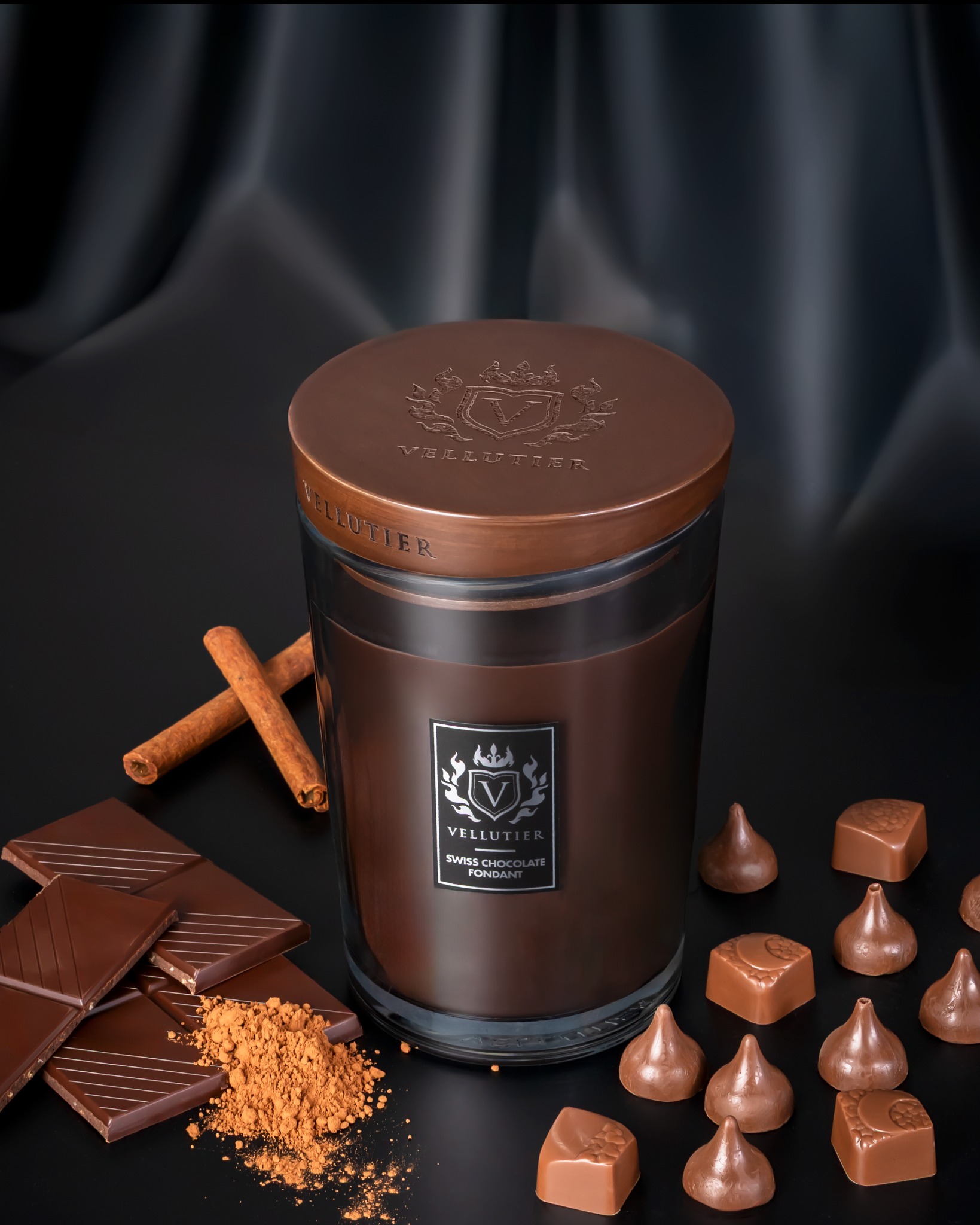 Fotografia produktowa: Świeca zapachowa Vellutier Swiss Chocolate Fondant na ciemnym tle, otoczona czekoladą, kakao i laskami cynamonu. Luksusowy charakter.