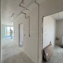 MK_TRO - Wnętrze w trakcie remontu: widoczne kable elektryczne prowadzone po ścianach i suficie, przygotowane do tynkowania. W tle osoba przy oknie i karton Geberit.