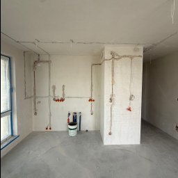 MK_TRO - Wnętrze w trakcie prac elektrycznych: kable i puszki podtynkowe rozprowadzone po ścianach, surowy beton na podłodze, widoczny narożnik i okno z niebieską taśmą.