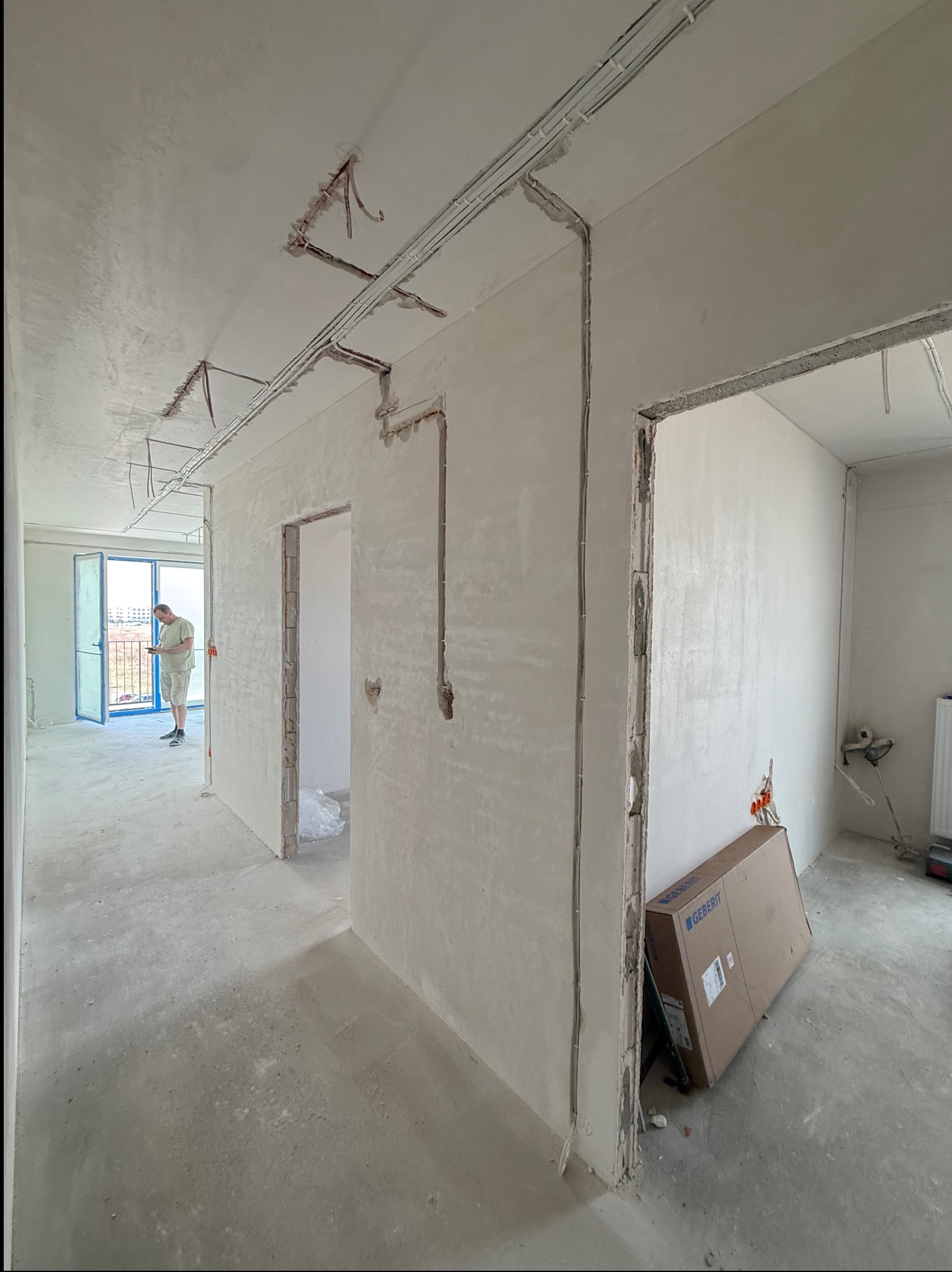 Wnętrze w trakcie remontu: widoczne kable elektryczne prowadzone po ścianach i suficie, przygotowane do tynkowania. W tle osoba przy oknie i karton Geberit.