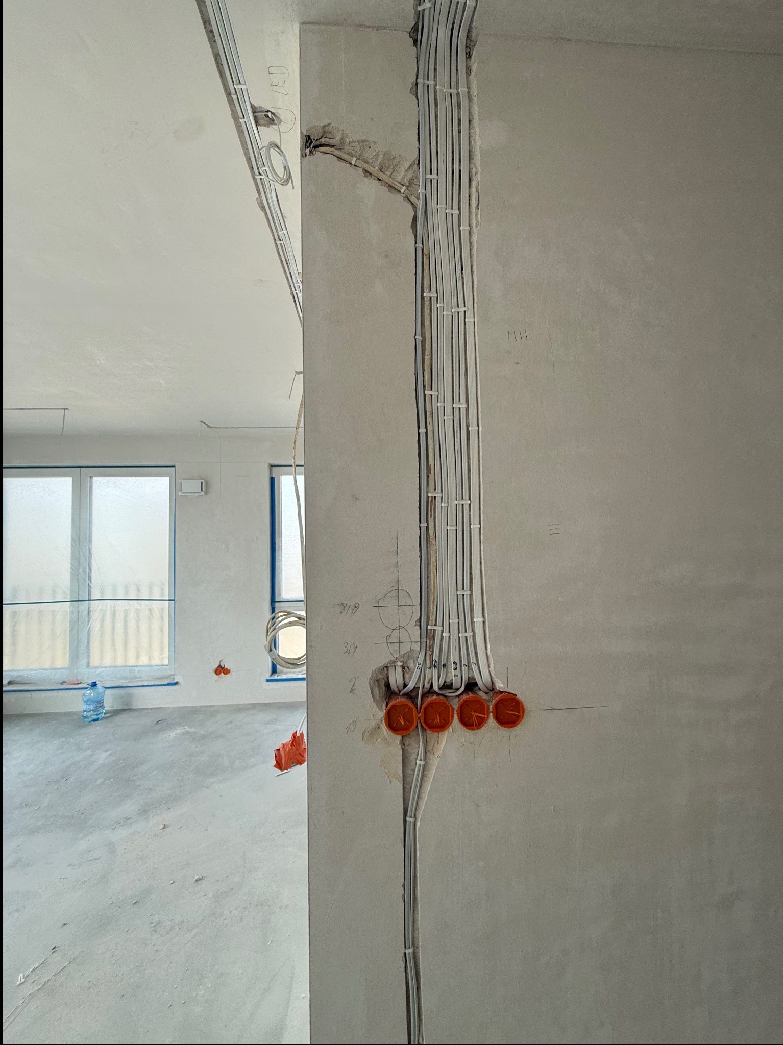 Białe kable elektryczne prowadzone wzdłuż ściany w surowym wnętrzu, z widocznymi pomarańczowymi puszkami instalacyjnymi i fragmentem okna w tle.