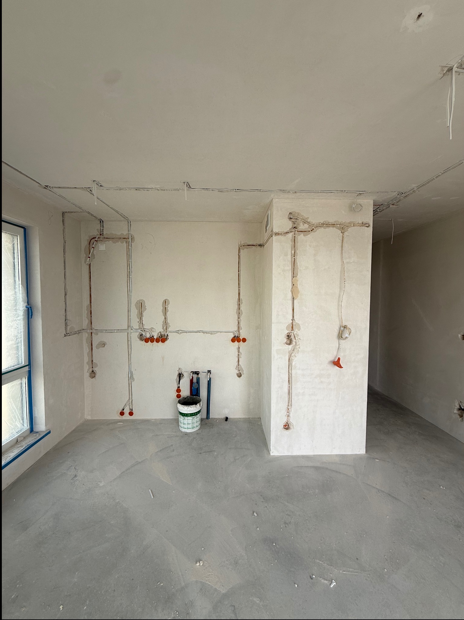 Wnętrze w trakcie prac elektrycznych: kable i puszki podtynkowe rozprowadzone po ścianach, surowy beton na podłodze, widoczny narożnik i okno z niebieską taśmą.