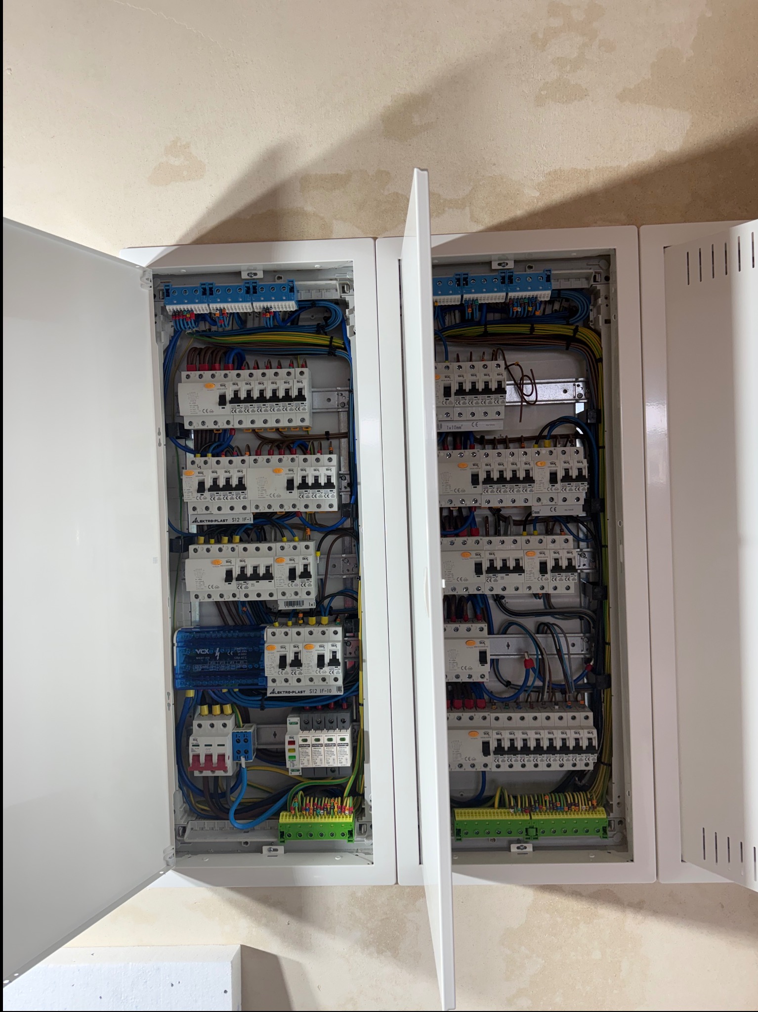 Otwarta, dwudrzwiowa, biała szafka rozdzielcza z widocznymi bezpiecznikami, przewodami i listwami zaciskowymi. Instalacja elektryczna wewnątrz budynku.