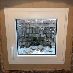 WOJPLAST - Białe okno piwniczne z widokiem na ceglany mur i gruz, świeżo osadzone w surowym murze. Widoczne ślady zaprawy i niedoskonałości ściany.