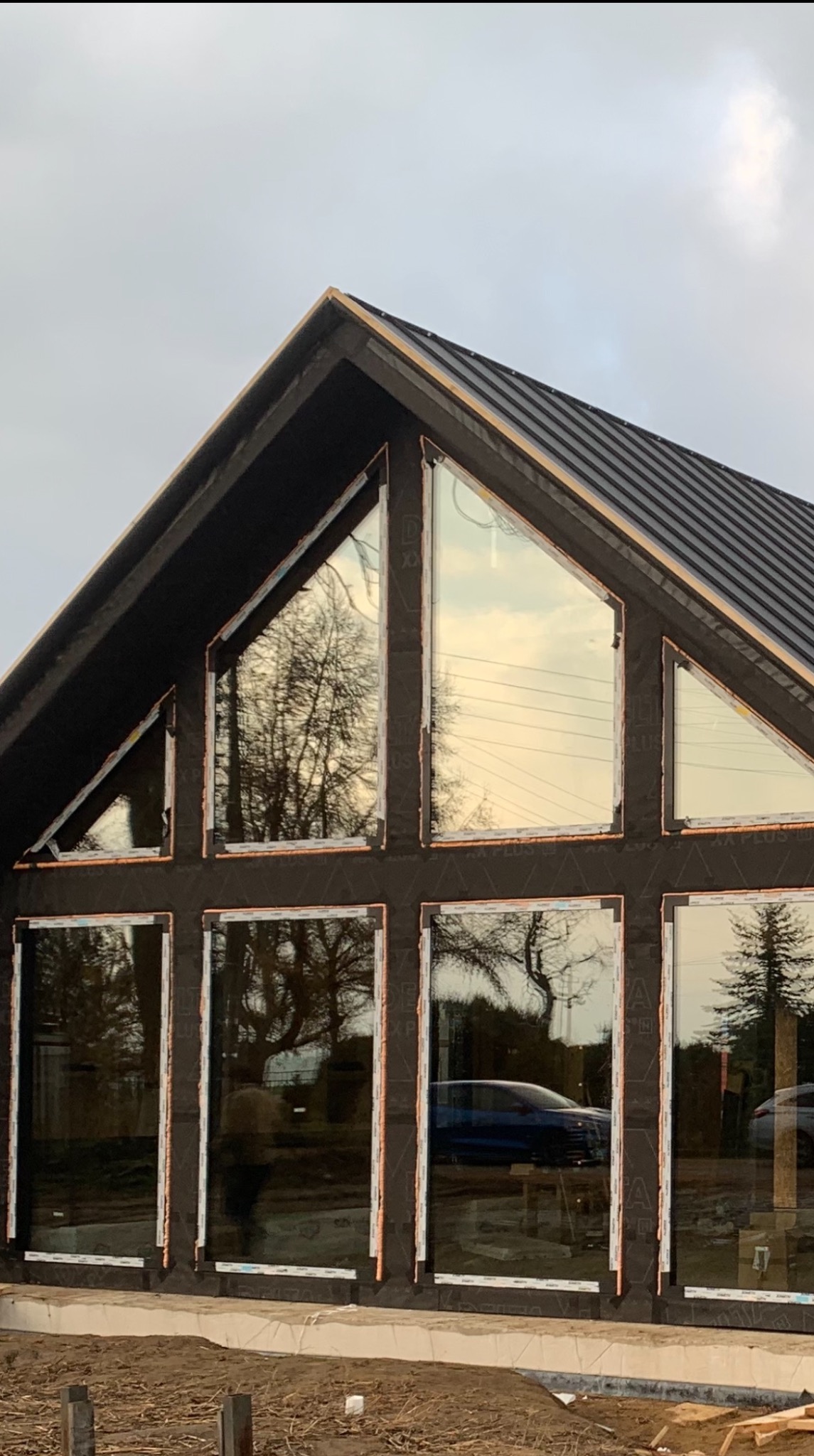 Nowoczesna fasada domu z dużymi oknami w trakcie montażu, widoczne zabezpieczenia. W szybach odbija się otoczenie, w tle pochmurne niebo.