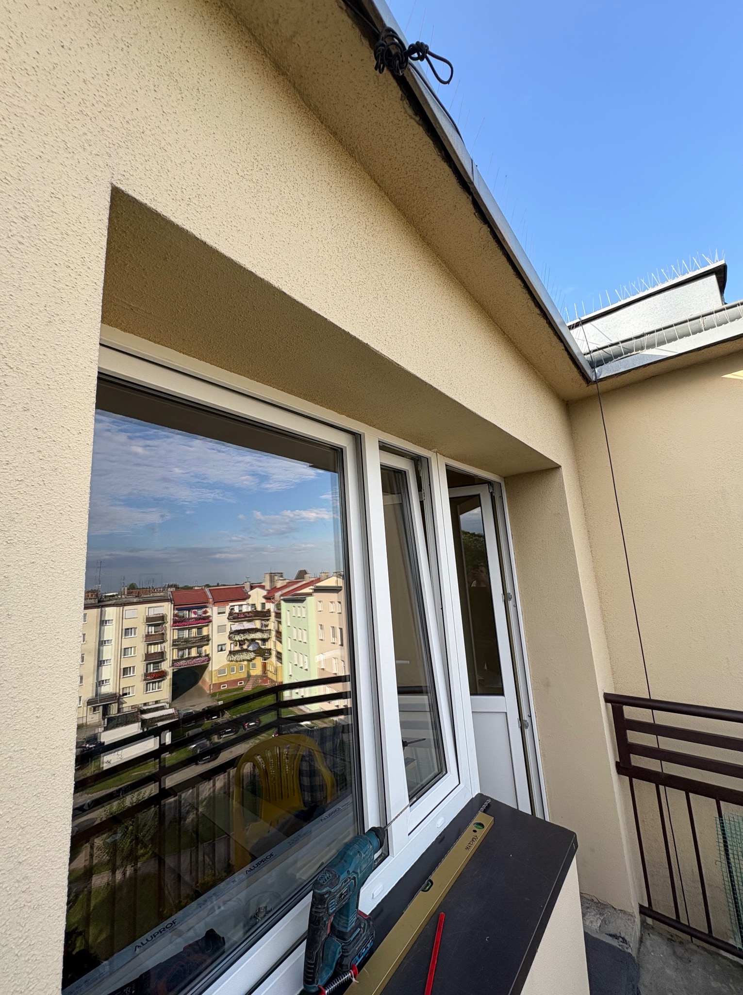 Białe okno PCV z widokiem na miasto, częściowo uchylone. Na parapecie poziomica i wiertarko-wkrętarka. Na dachu kolce przeciw ptakom. Jasne, naturalne światło.