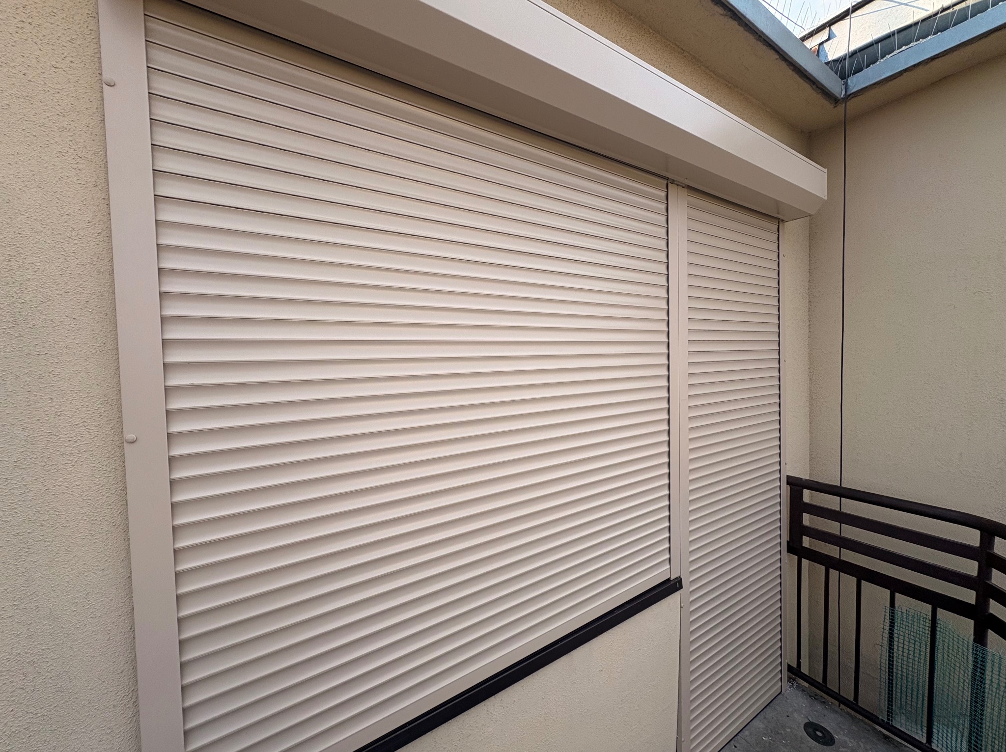 Zamknięte, beżowe rolety zewnętrzne na balkonie, widoczne z bliska, z fragmentem balustrady. Ochrona przed słońcem i wiatrem, zapewniają prywatność.