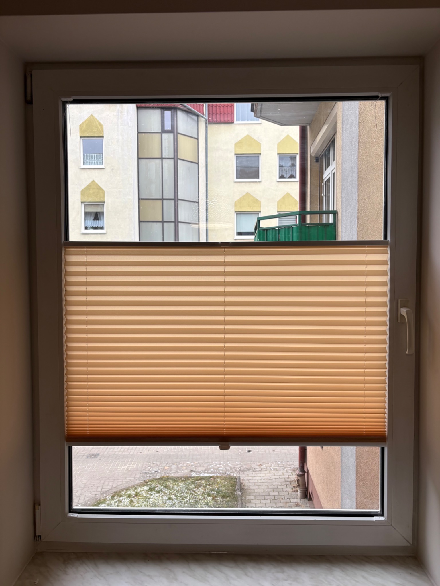 Okno PCV z beżową plisą okienną, widok na budynek z zielonym balkonem. Plisa częściowo zasłonięta, widoczny fragment parapetu z marmurkowym wzorem.