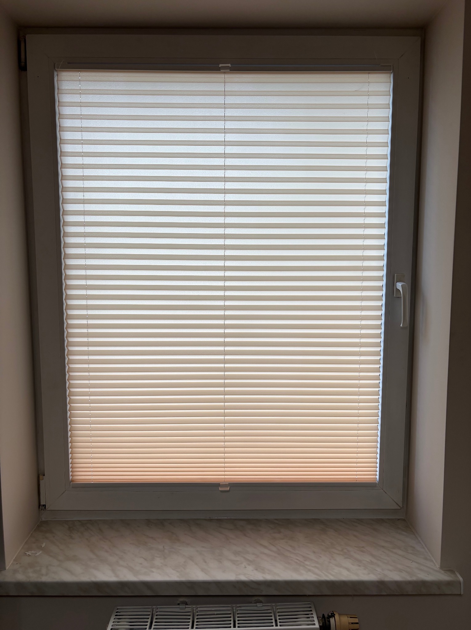 Okno PCV z plisowanymi roletami w Głubczycach. Biała rama okienna, marmurowy parapet. Widoczne żebra grzejnika pod oknem. Delikatne światło wpadające przez rolety.