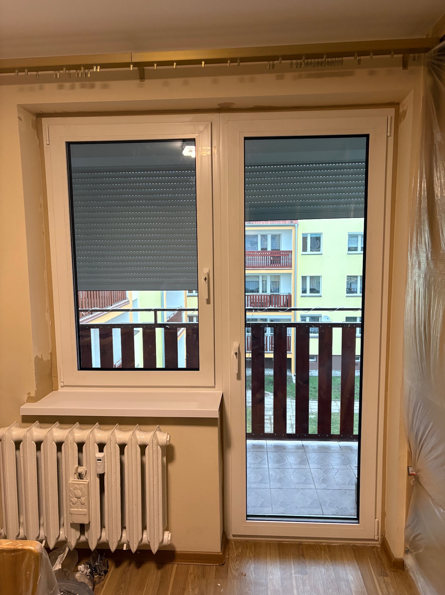 Białe okno balkonowe z roletami wewnętrznymi, widok na balkon i budynek. Grzejnik pod oknem, ściany przygotowane do malowania. Ujęcie z wnętrza mieszkania.