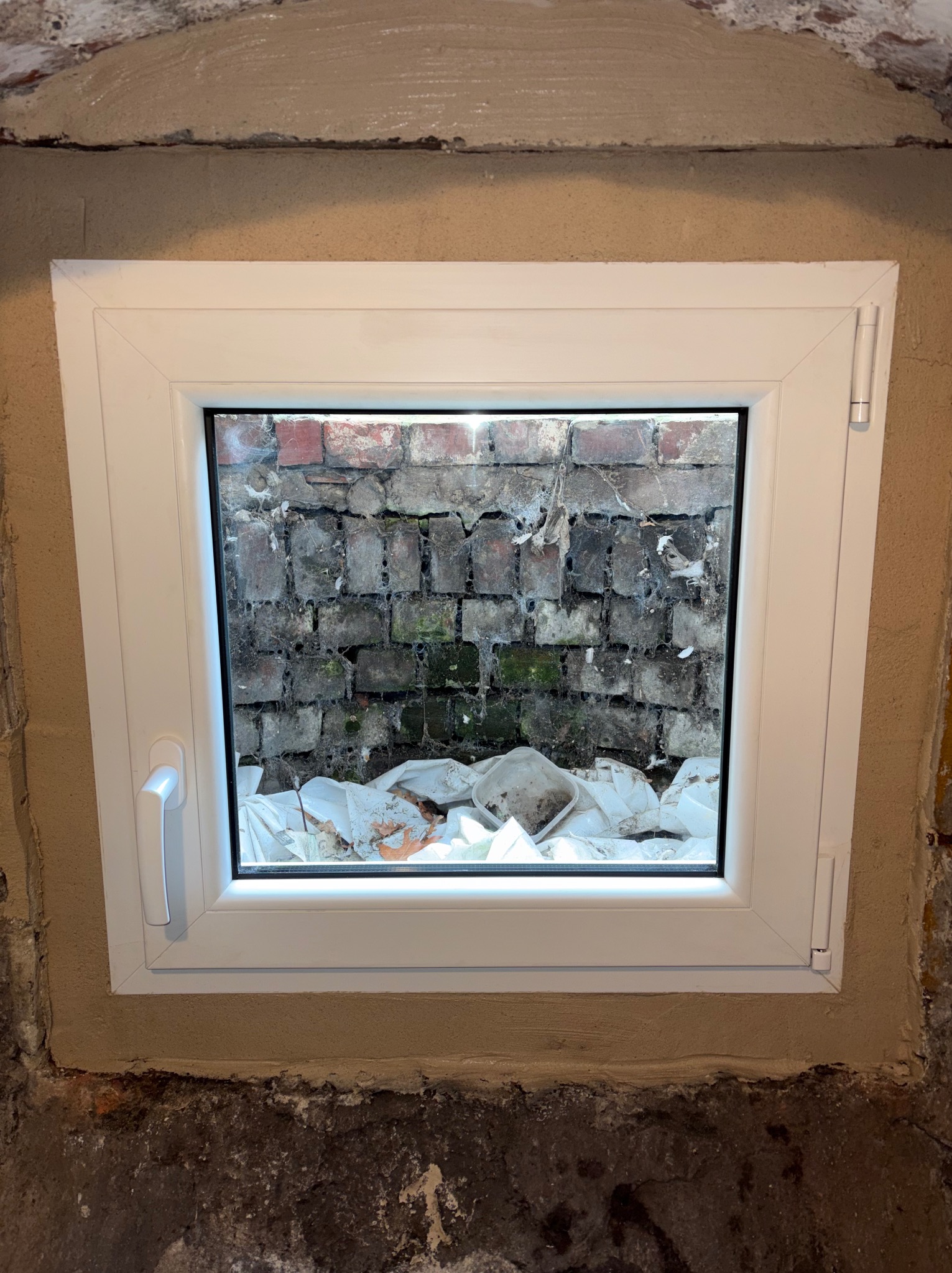 Białe okno piwniczne z widokiem na ceglany mur i gruz, świeżo osadzone w surowym murze. Widoczne ślady zaprawy i niedoskonałości ściany.