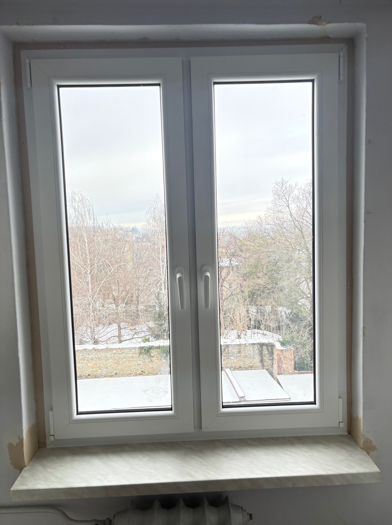 Białe, dwuskrzydłowe okno PCV z widokiem na zimowy krajobraz. Widoczne ślady po montażu na ścianie i parapet z imitacją marmuru. Grzejnik pod parapetem.