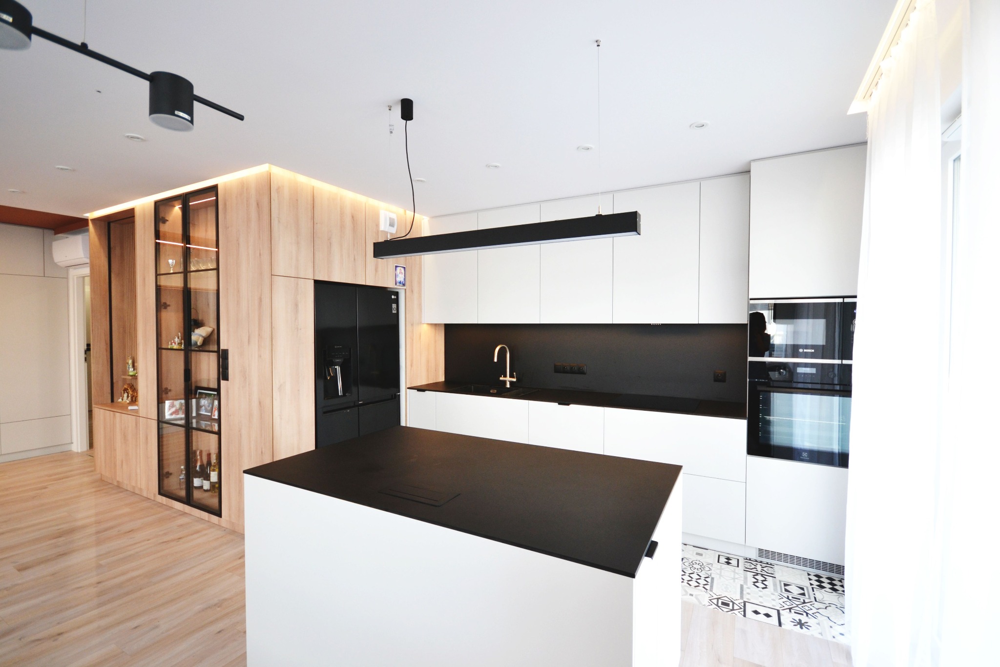 Nowoczesna kuchnia z wyspą, białe szafki, czarny blat i sprzęt AGD. Drewniana zabudowa z podświetleniem. Dekoracyjne płytki podłogowe.