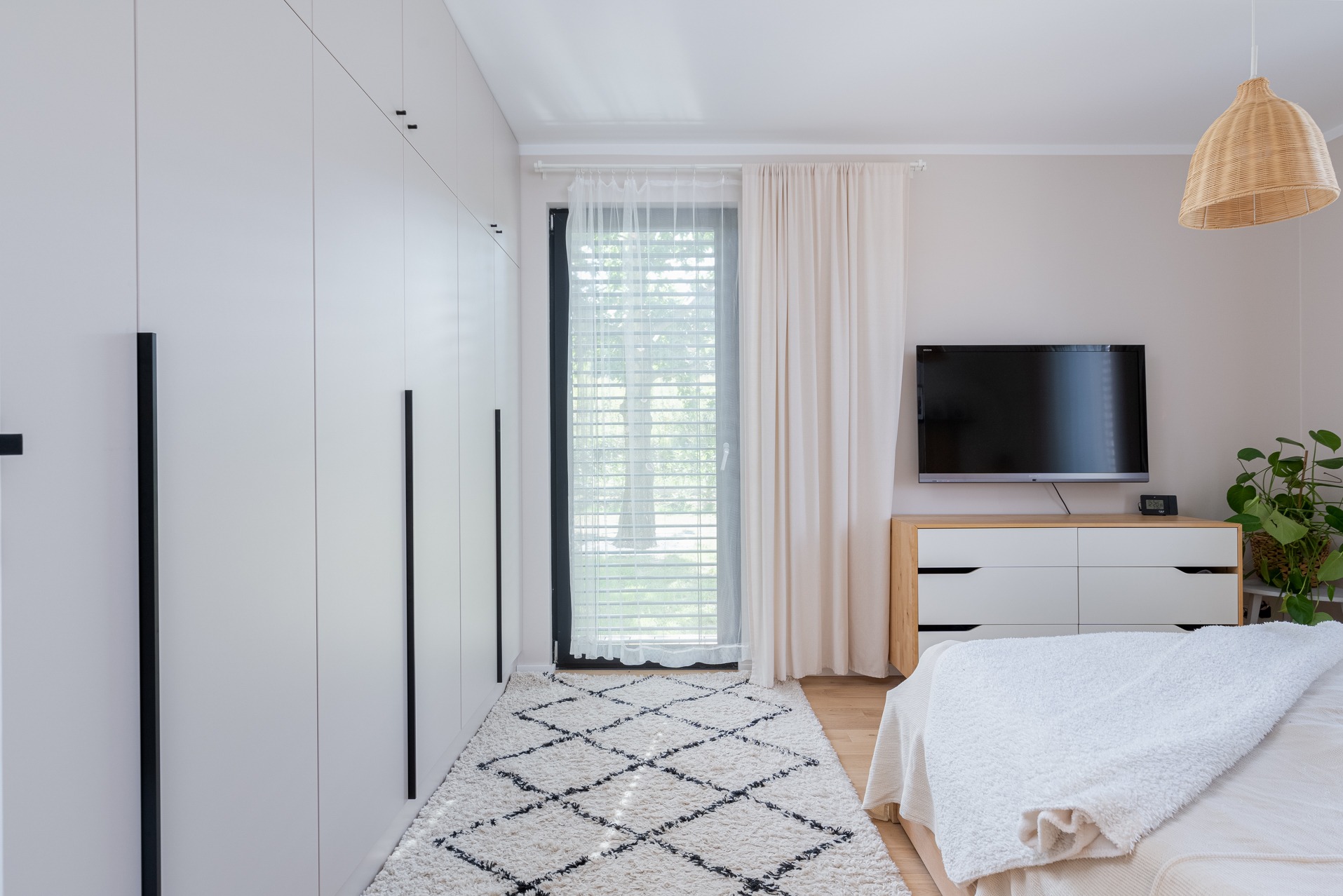 Nowoczesna sypialnia z białymi szafami na wymiar z czarnymi uchwytami, dywanem w romby i komodą pod telewizorem. Minimalistyczny design wnętrza.