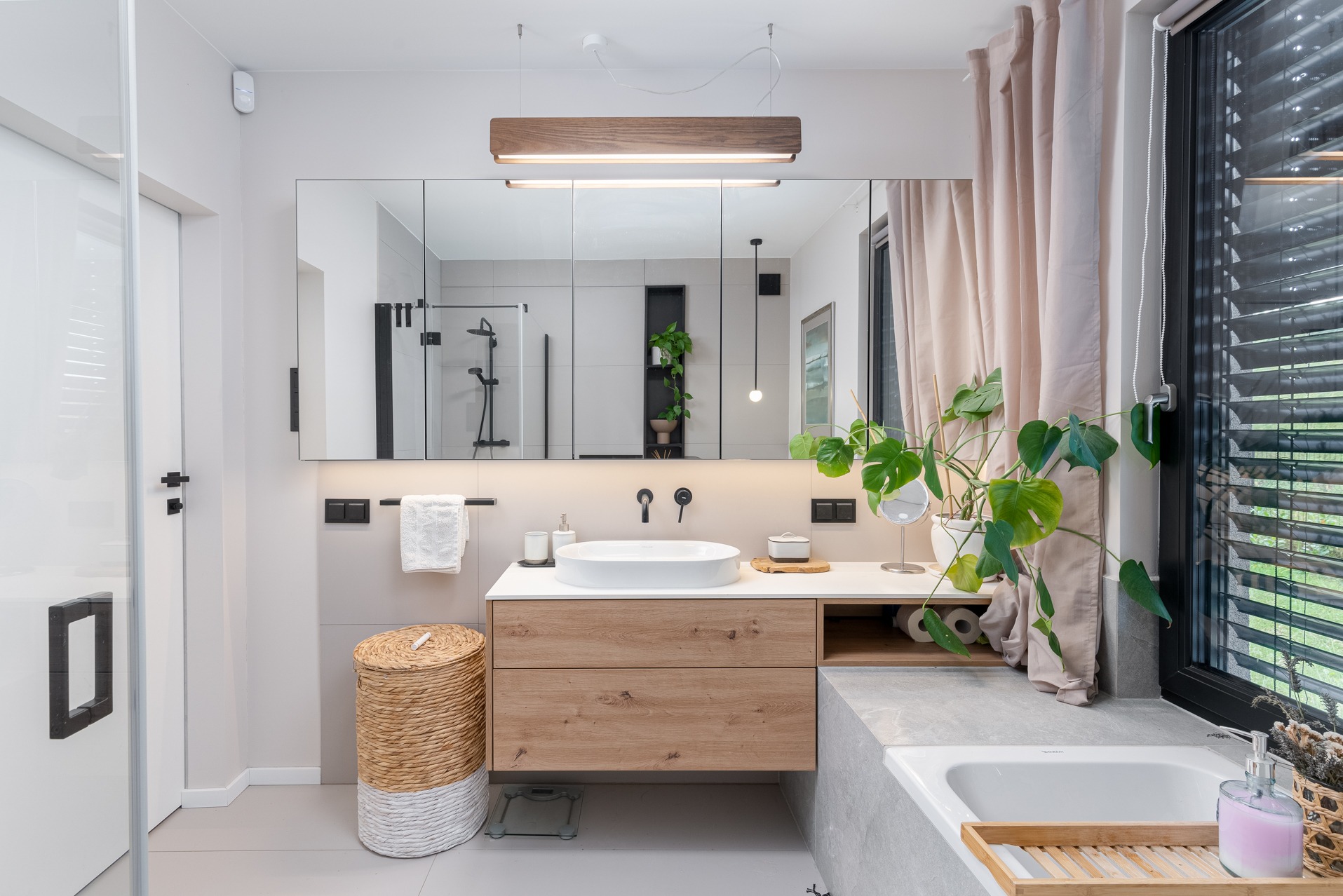 Nowoczesna łazienka z drewnianą szafką pod umywalką, dużym lustrem i roślinami. Minimalistyczny design, jasne kolory, naturalne materiały i oświetlenie LED.