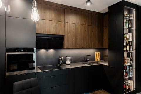 Nowoczesna kuchnia z ciemnymi szafkami i drewnianymi akcentami, oświetleniem LED oraz przeszkloną witryną z alkoholem. Minimalistyczny design z wiszącą lampą.