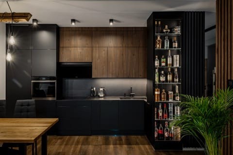 Nowoczesna kuchnia z czarnymi frontami i drewnianymi akcentami, oświetleniem punktowym i przeszkloną witryną z alkoholem. Minimalistyczny design z rośliną doniczkową.