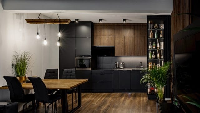 Nowoczesna kuchnia z czarnymi frontami i drewnianymi akcentami. Na blacie czajnik i oświetlenie LED. Stół z krzesłami i dekoracyjna lampa wisząca nad nim.