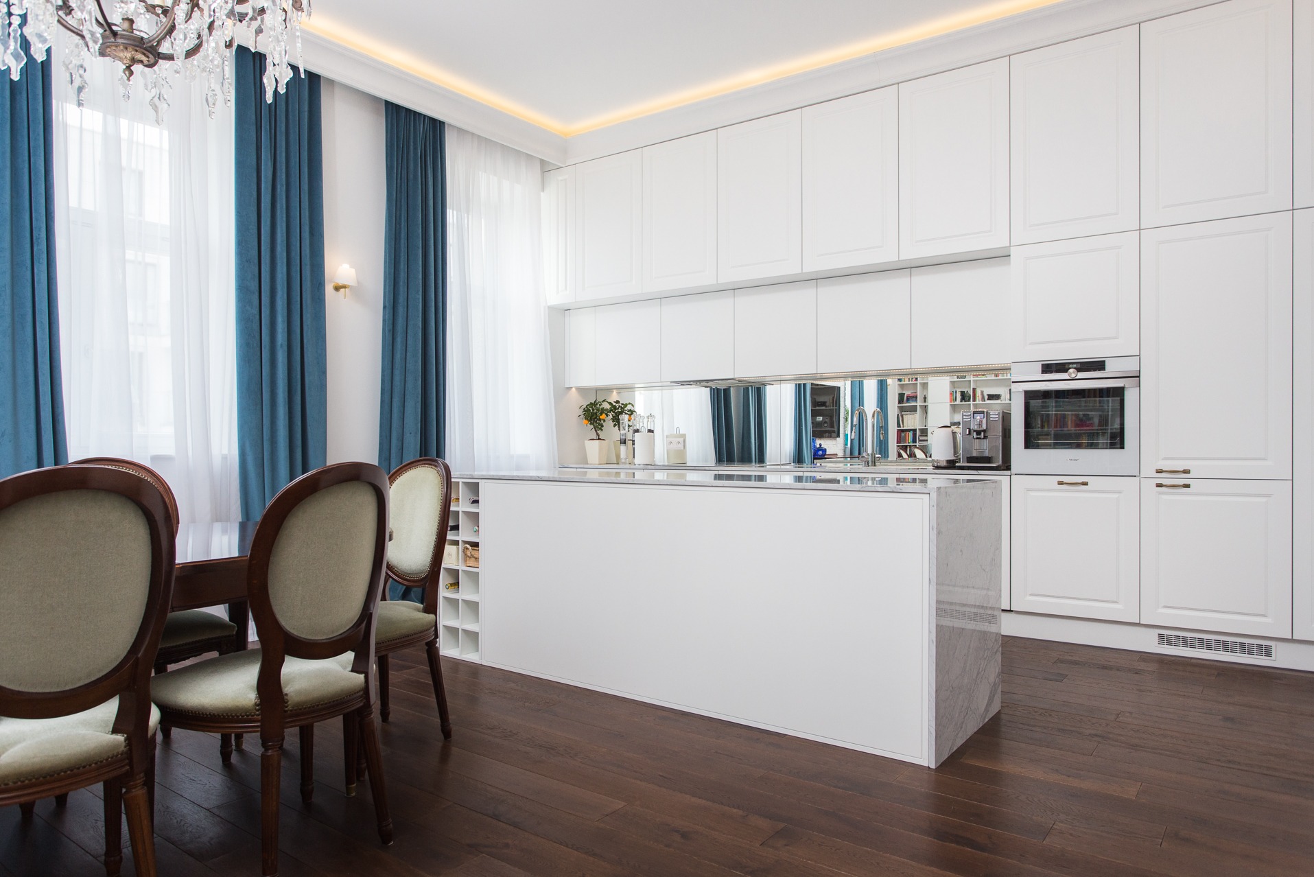 Nowoczesna kuchnia z białą zabudową i wyspą z marmurowym blatem. W tle okno z turkusowymi zasłonami, drewniana podłoga i stół z krzesłami.