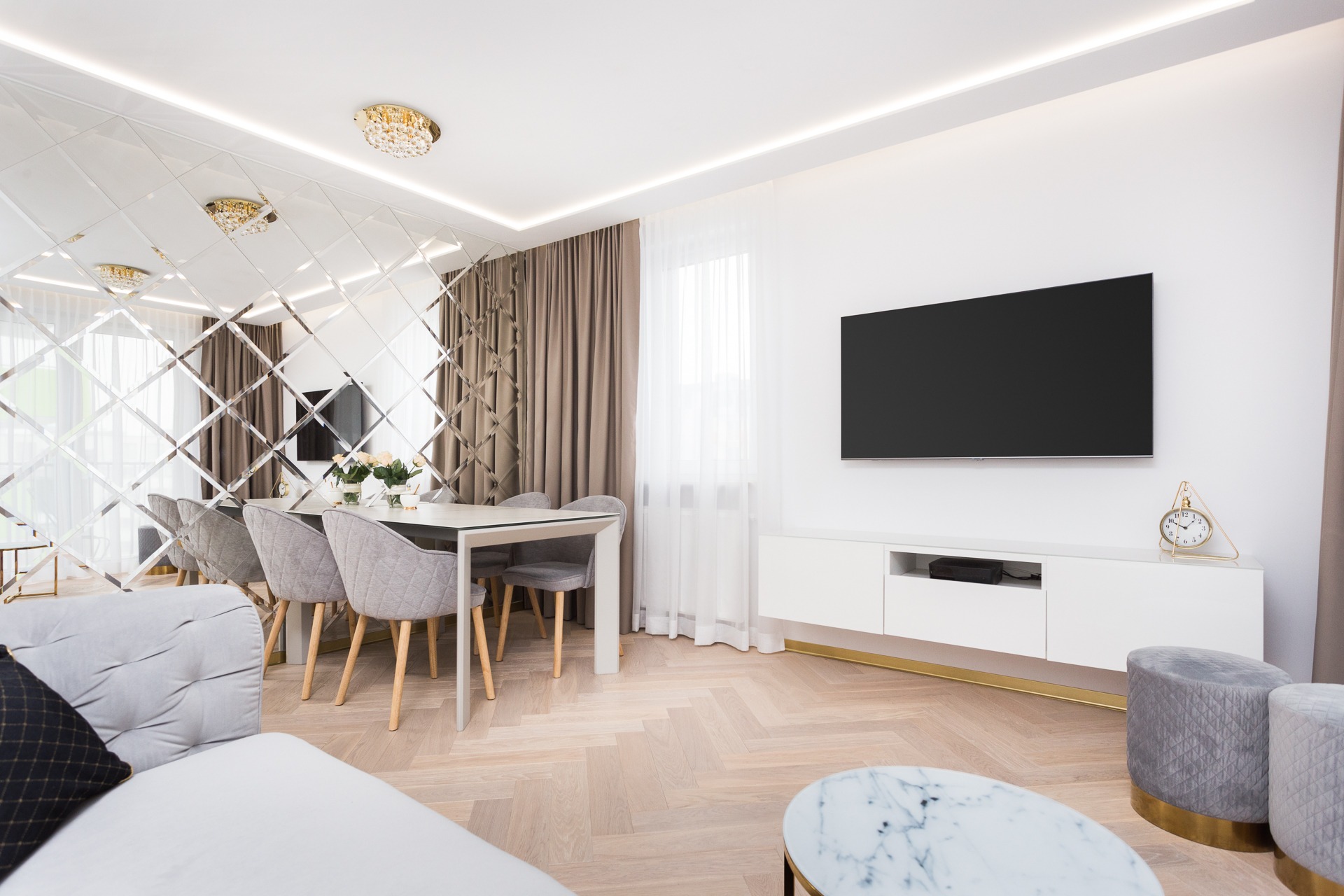 Elegancka, jasna jadalnia z lustrzaną ścianą, białym stołem i szarymi krzesłami. Na ścianie telewizor, a na komodzie złoty zegar. Parkiet ułożony w jodełkę.