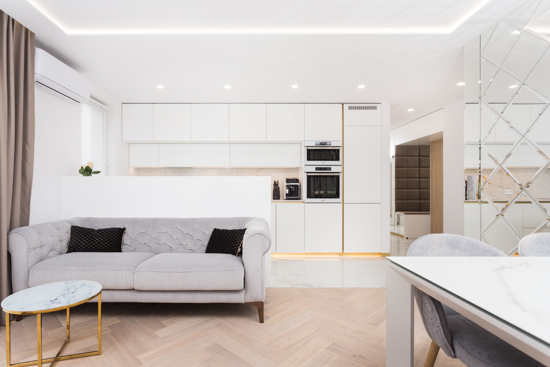 Elegancka, otwarta przestrzeń: biała zabudowa kuchenna z piekarnikiem i mikrofalówką, szara sofa z poduszkami i stolikiem na tle parkietu w jodełkę.