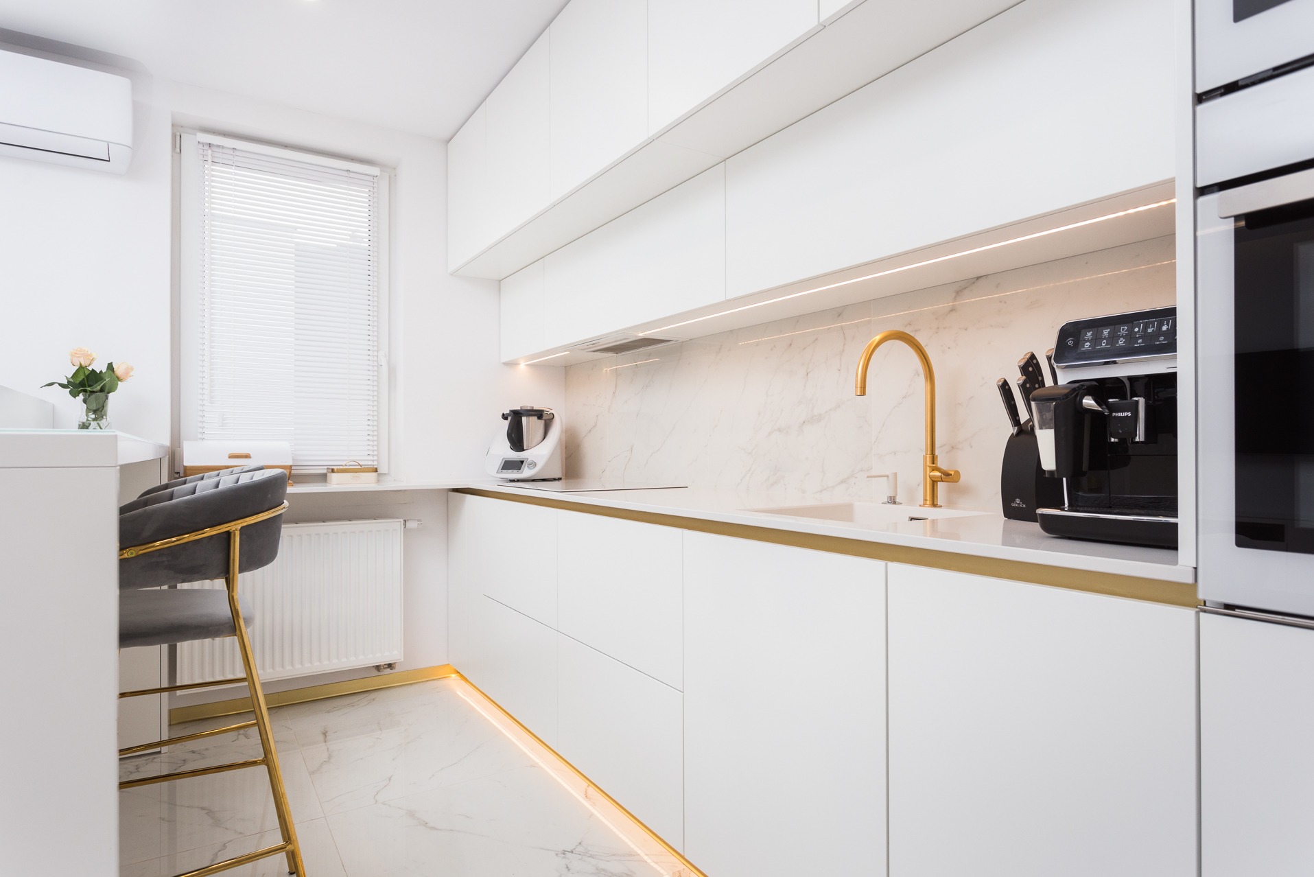 Nowoczesna, biała kuchnia z marmurowym backsplashiem i złotymi akcentami, oświetleniem LED, sprzętem AGD i krzesłem barowym. Minimalistyczny design.