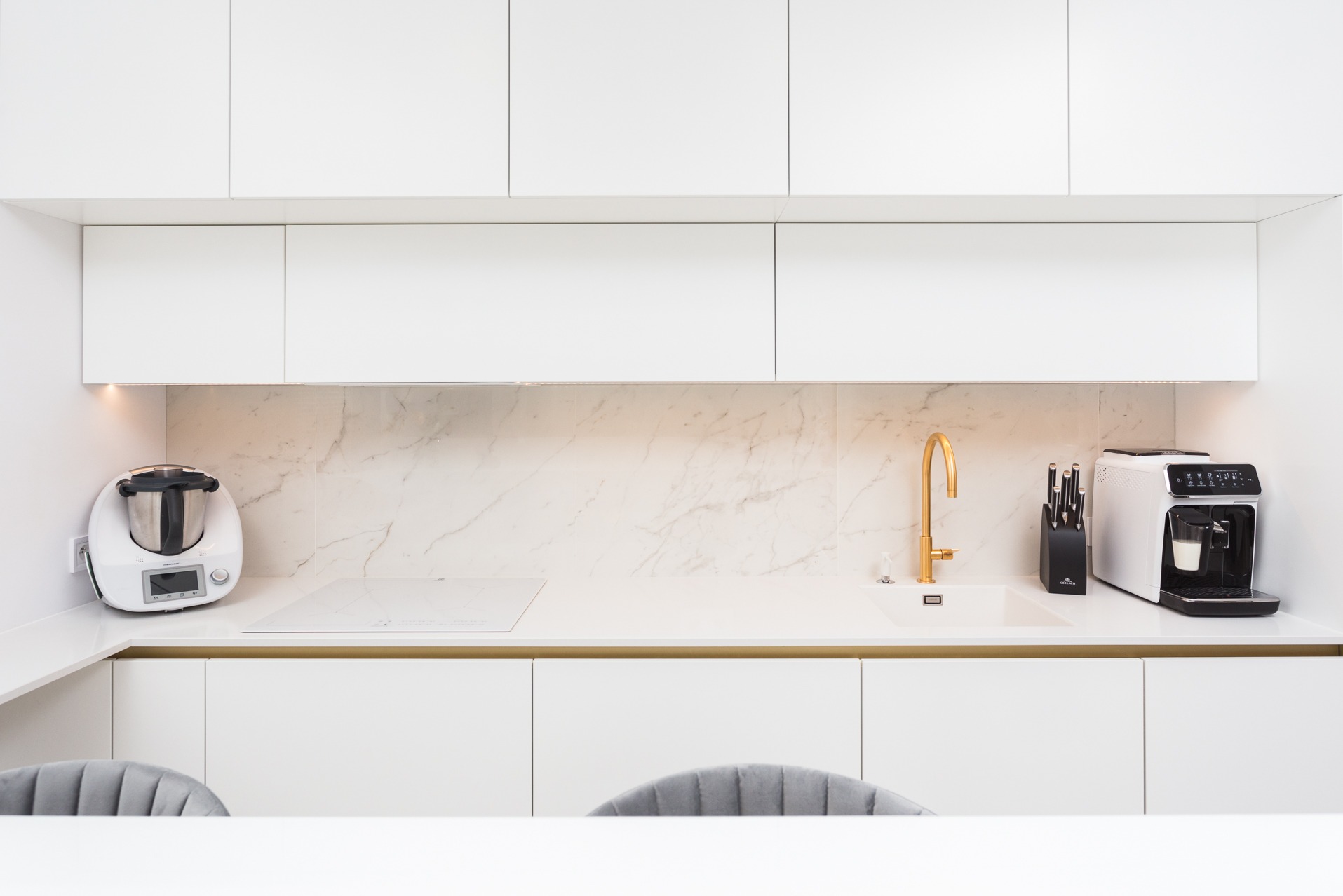Biała kuchnia z marmurowym blatem, złotą armaturą i sprzętami AGD: Thermomix, płyta indukcyjna, ekspres do kawy. Minimalistyczny design.