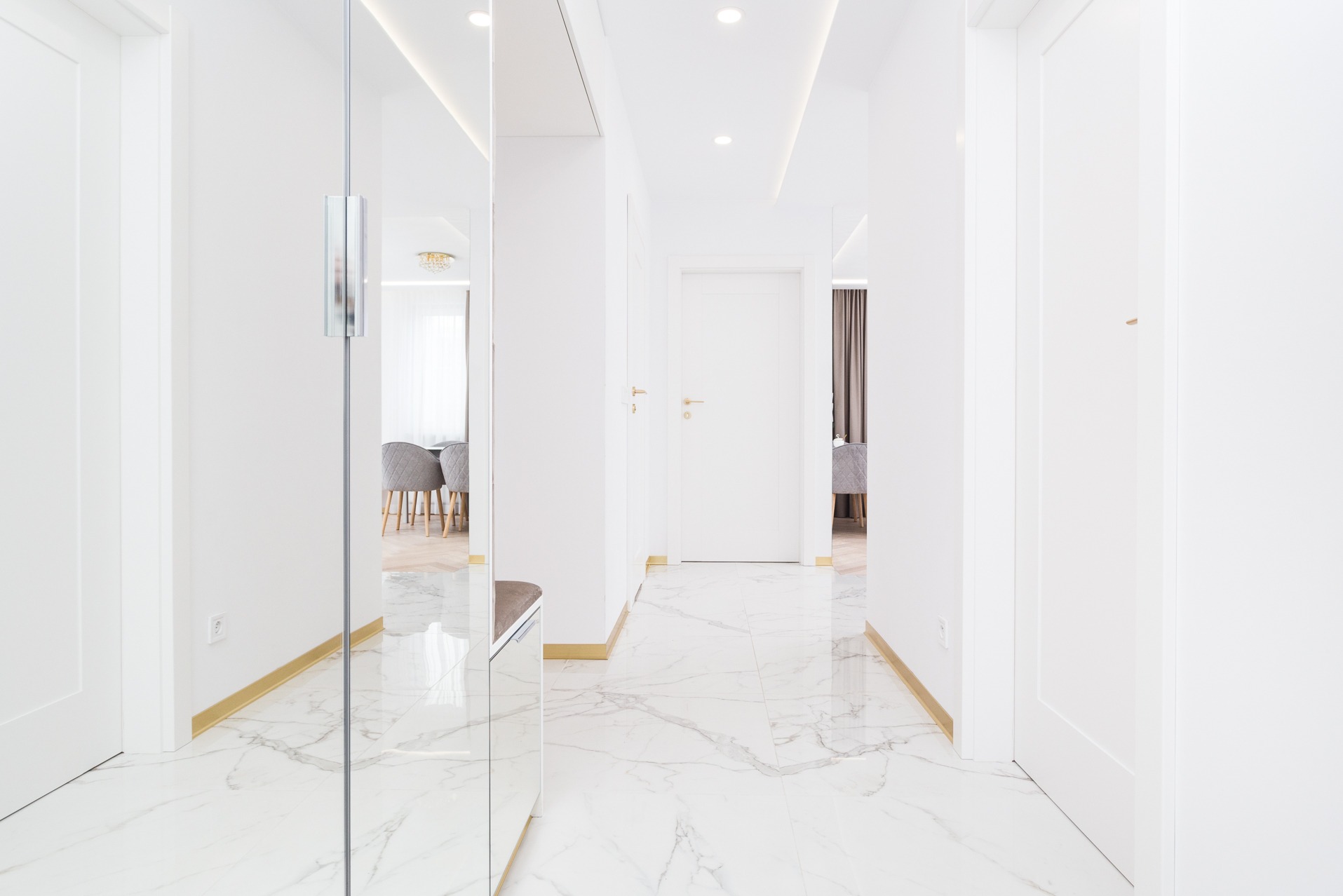 Białe wnętrze przedpokoju z marmurową podłogą i lustrem w zabudowie. Widok na jadalnię z szarymi krzesłami. Minimalistyczny design, złote akcenty.