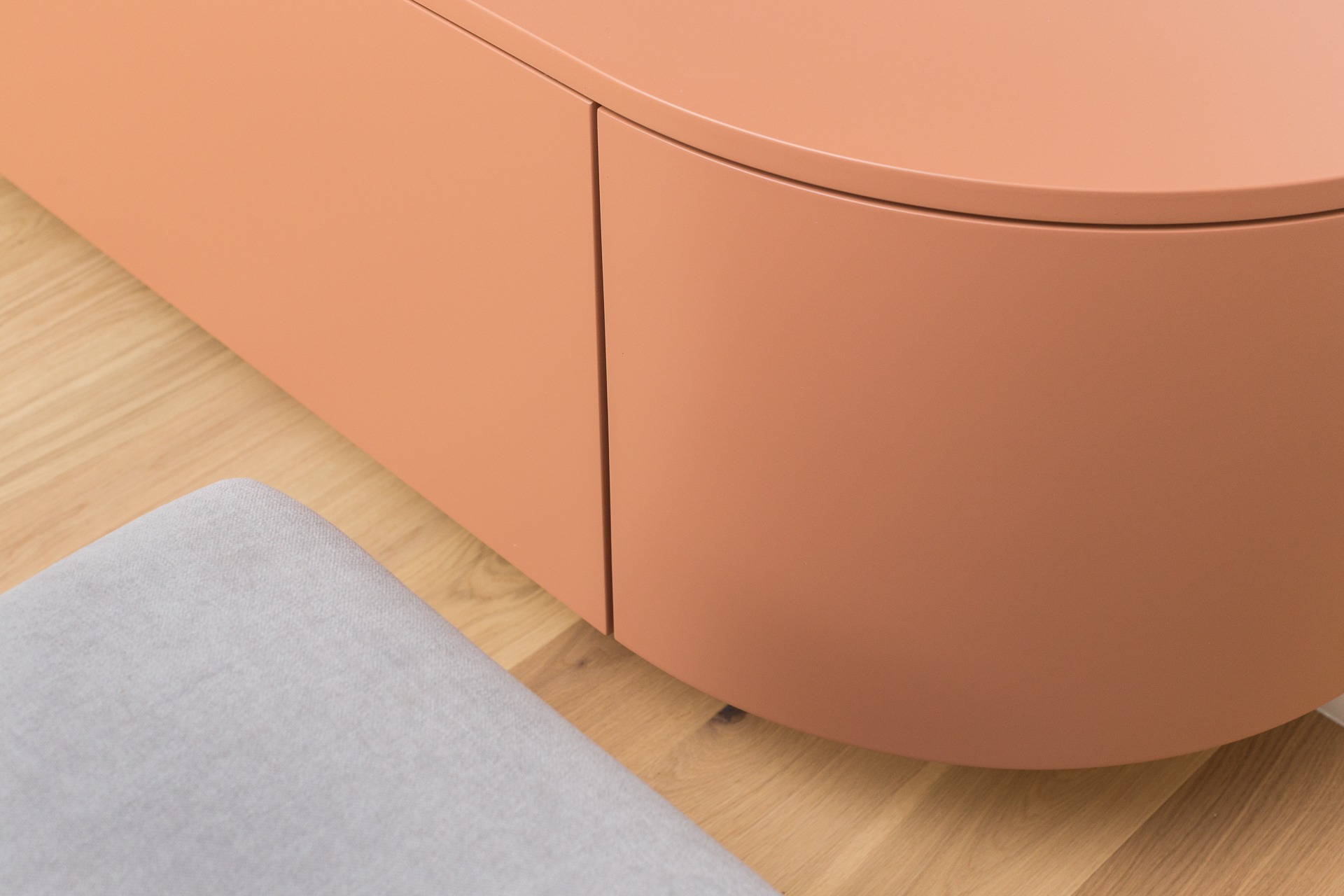 Zaokrąglona szafka w odcieniu brzoskwini, z szarą pufą obok, na drewnianej podłodze. Minimalistyczny design, gładkie powierzchnie i subtelne oświetlenie.