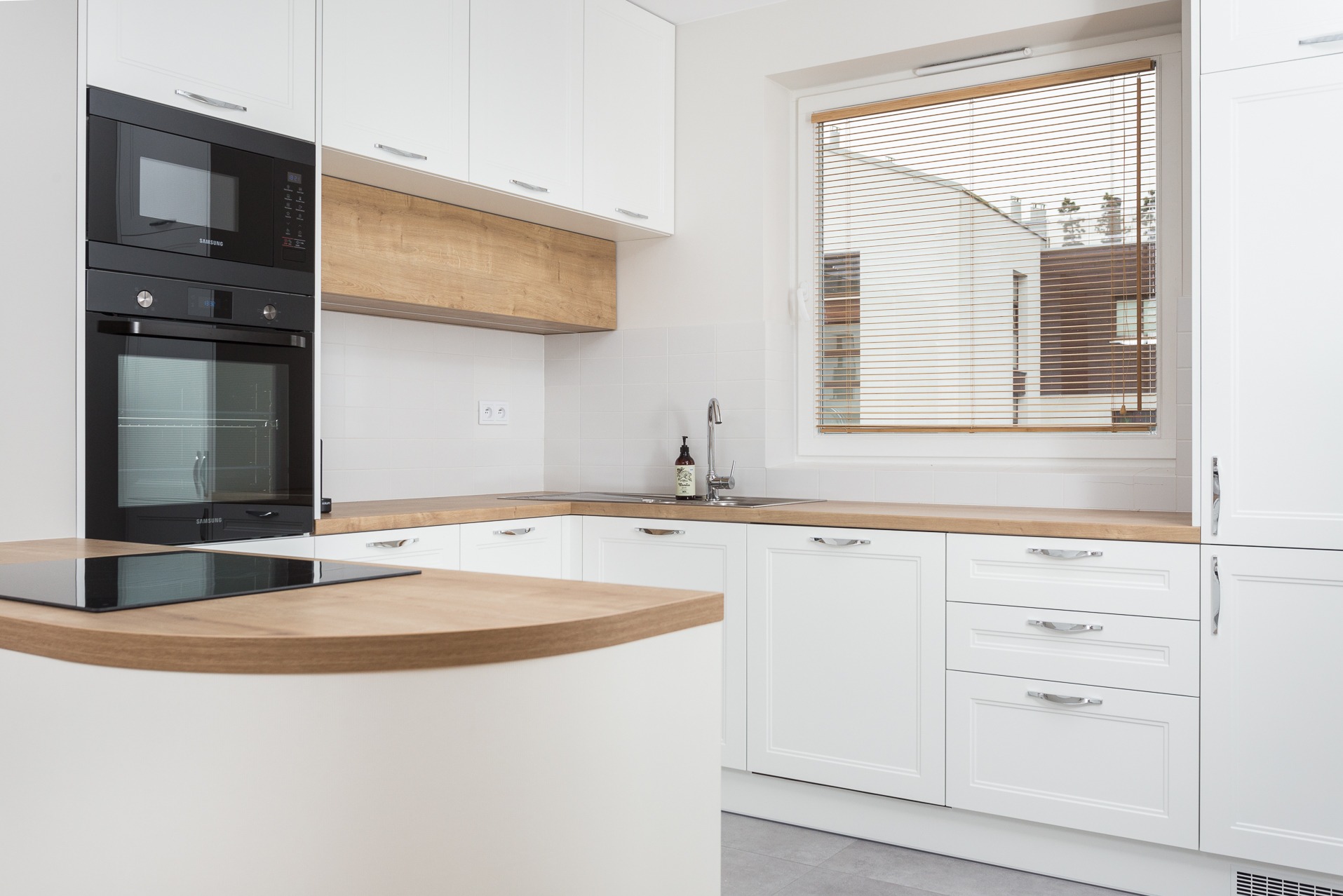 Jasna kuchnia z białymi szafkami, drewnianym blatem i sprzętem Samsung. Widoczne okno z żaluzjami i minimalistyczny design. Nowoczesny styl.