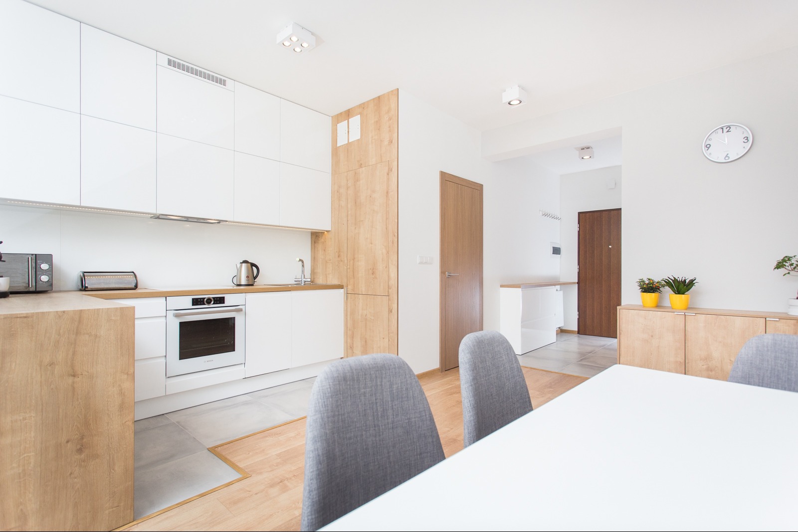 Jasna kuchnia z białymi szafkami i drewnianymi akcentami, widok z jadalni. Nowoczesny design, minimalistyczne oświetlenie, szare krzesła przy białym stole.