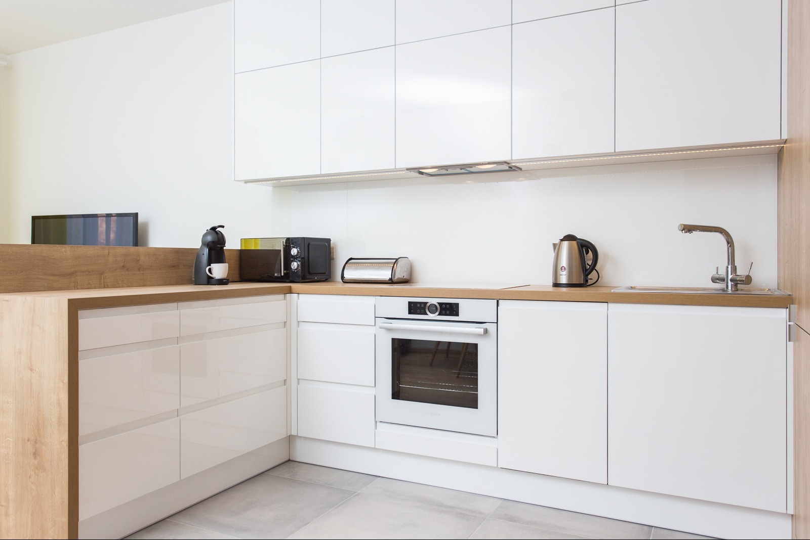 Nowoczesna, jasna kuchnia z białymi, lakierowanymi frontami, drewnianym blatem i sprzętami AGD: piekarnik, czajnik, mikrofalówka, ekspres do kawy.