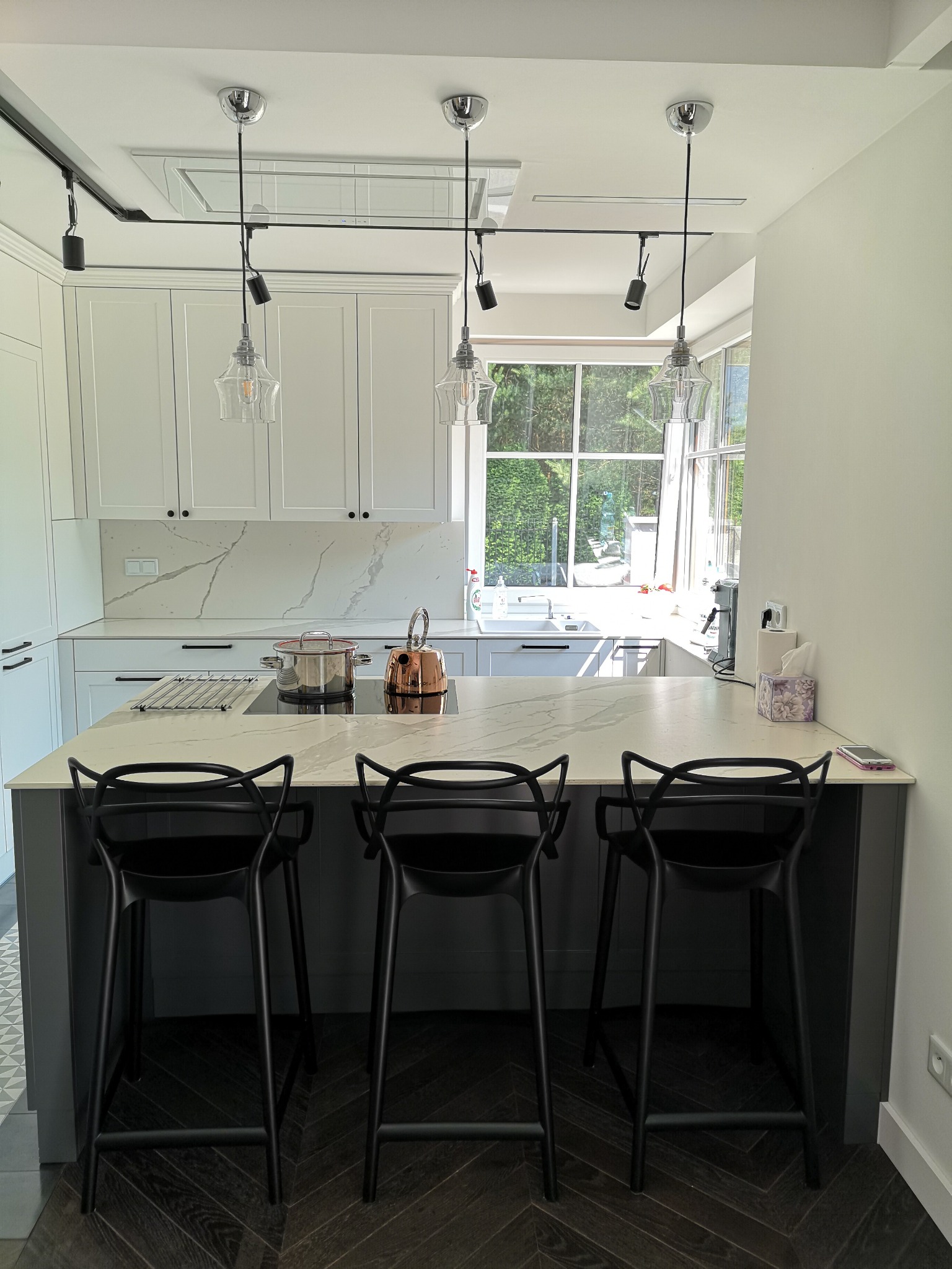 Biała kuchnia z wyspą i trzema czarnymi hokerami. Na blacie marmurowy wzór, okno z widokiem na zieleń. Nad wyspą wiszące lampy. Nowoczesny styl.