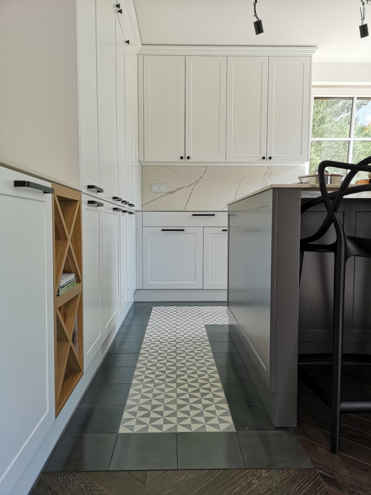 Nowoczesna kuchnia z białymi szafkami, czarnymi uchwytami i wzorzystą podłogą. W tle okno z widokiem na zieleń. Widoczny fragment wyspy kuchennej i krzesło barowe.