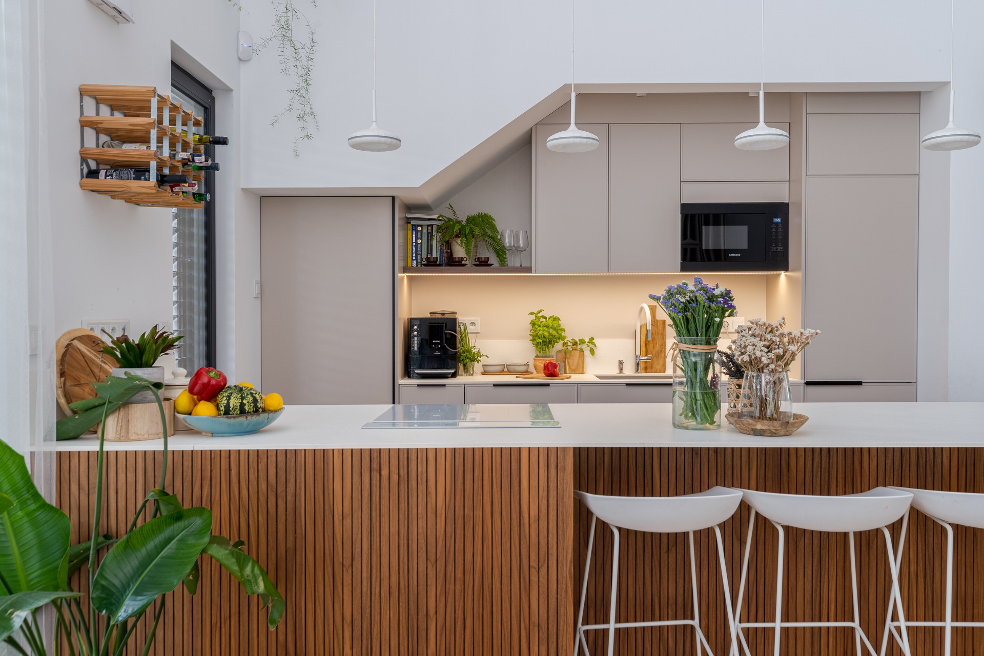 Jasna kuchnia z drewnianym barkiem i białymi hokerami. Na blacie owoce, kwiaty w wazonach, zlew i ekspres do kawy. Na ścianie półka na wino i wiszące rośliny.