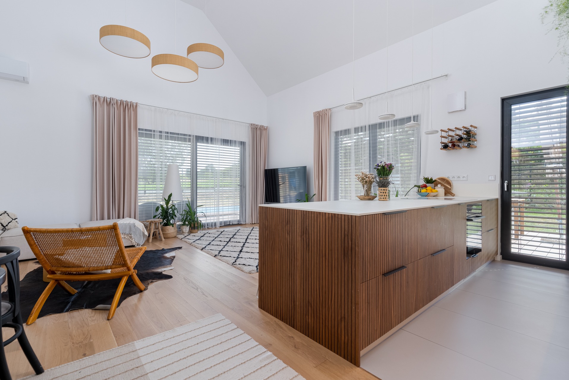 Jasne wnętrze z drewnianą wyspą kuchenną i widokiem na ogród przez duże okna. Nowoczesne oświetlenie i minimalistyczny design. Elementy dekoracyjne dodają przytulności.