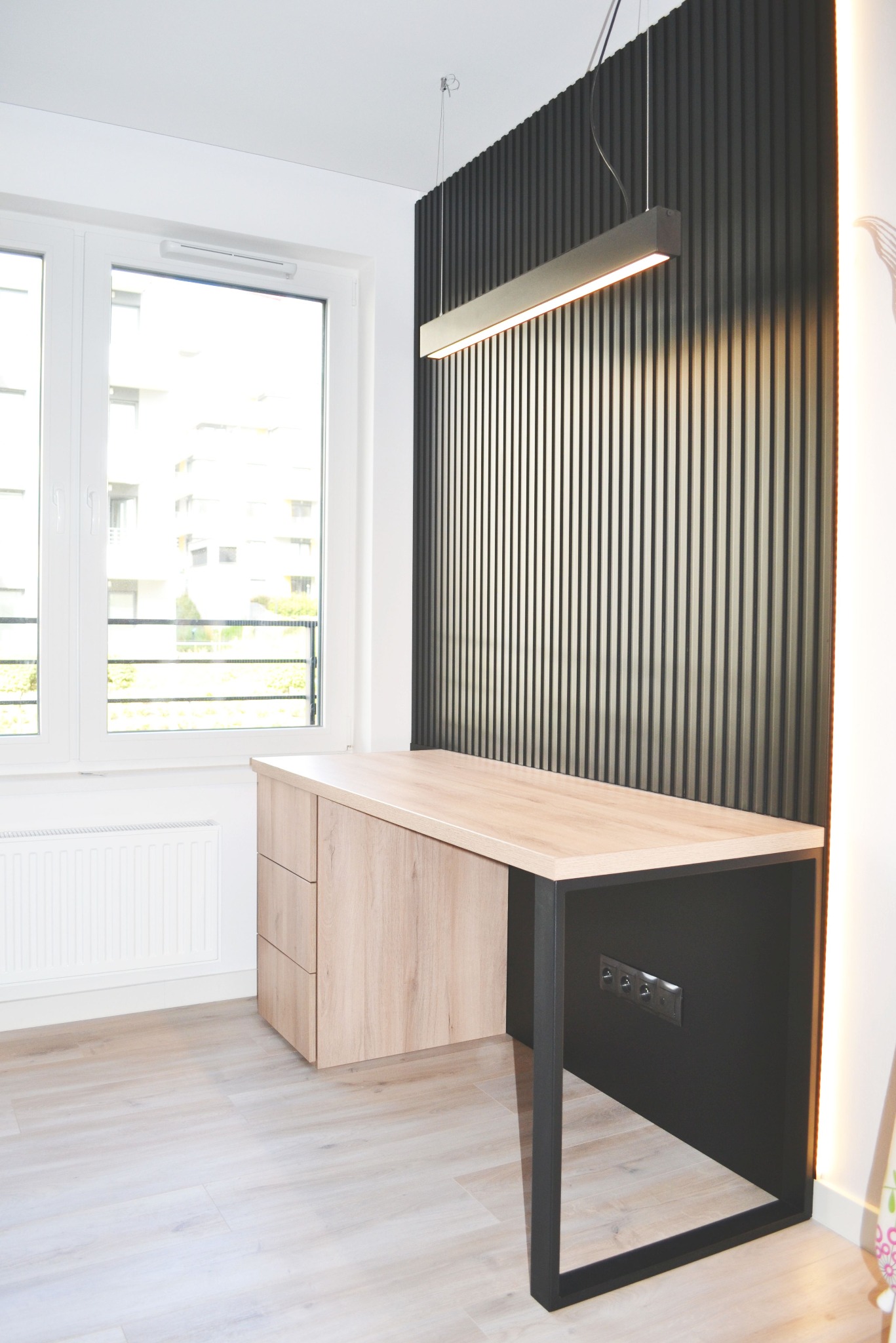 Nowoczesne biurko na wymiar z drewnianym blatem i czarnymi nogami, zintegrowane gniazdka, oświetlenie ledowe, na tle ściany z pionowych paneli dekoracyjnych.