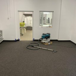 Firma Budowlana TOM-BUD TOMASZ PRZYCHODZEŃ - Szary dywan w pustym pomieszczeniu po wykończeniu, z odkurzaczem przemysłowym Makita. W tle widoczne białe ściany, okno i otwarte drzwi.