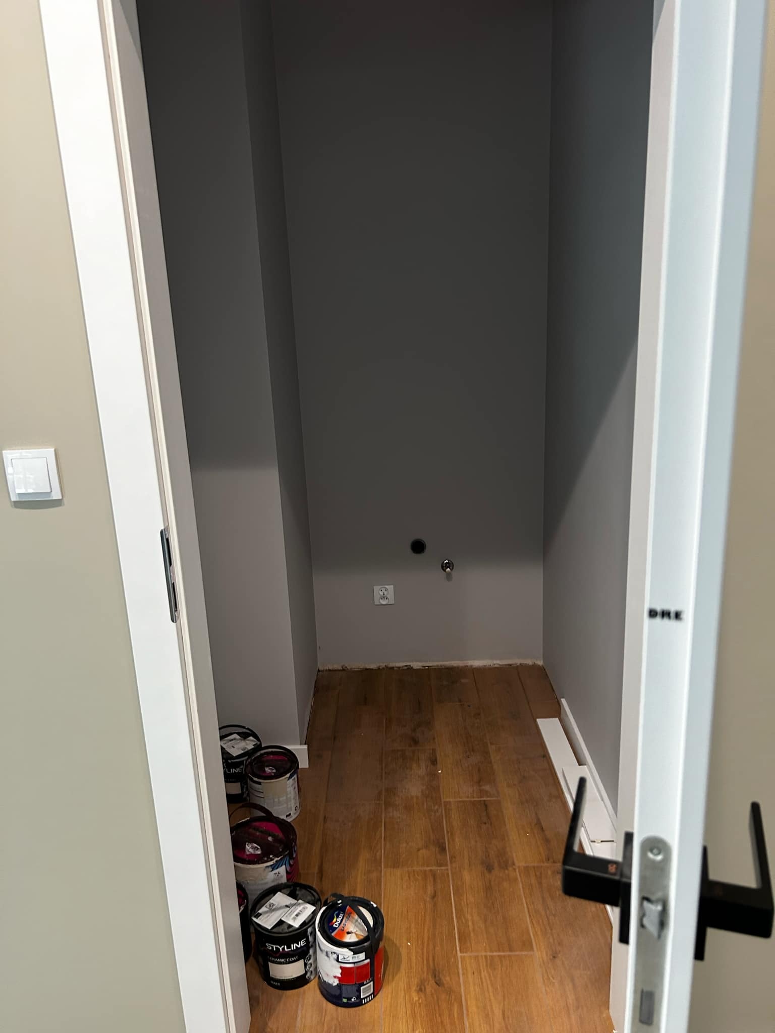 Wnętrze małego pomieszczenia z drewnianą podłogą i szarymi ścianami, widoczne puszki z farbą przy wejściu. Otwór drzwiowy z białą ościeżnicą.