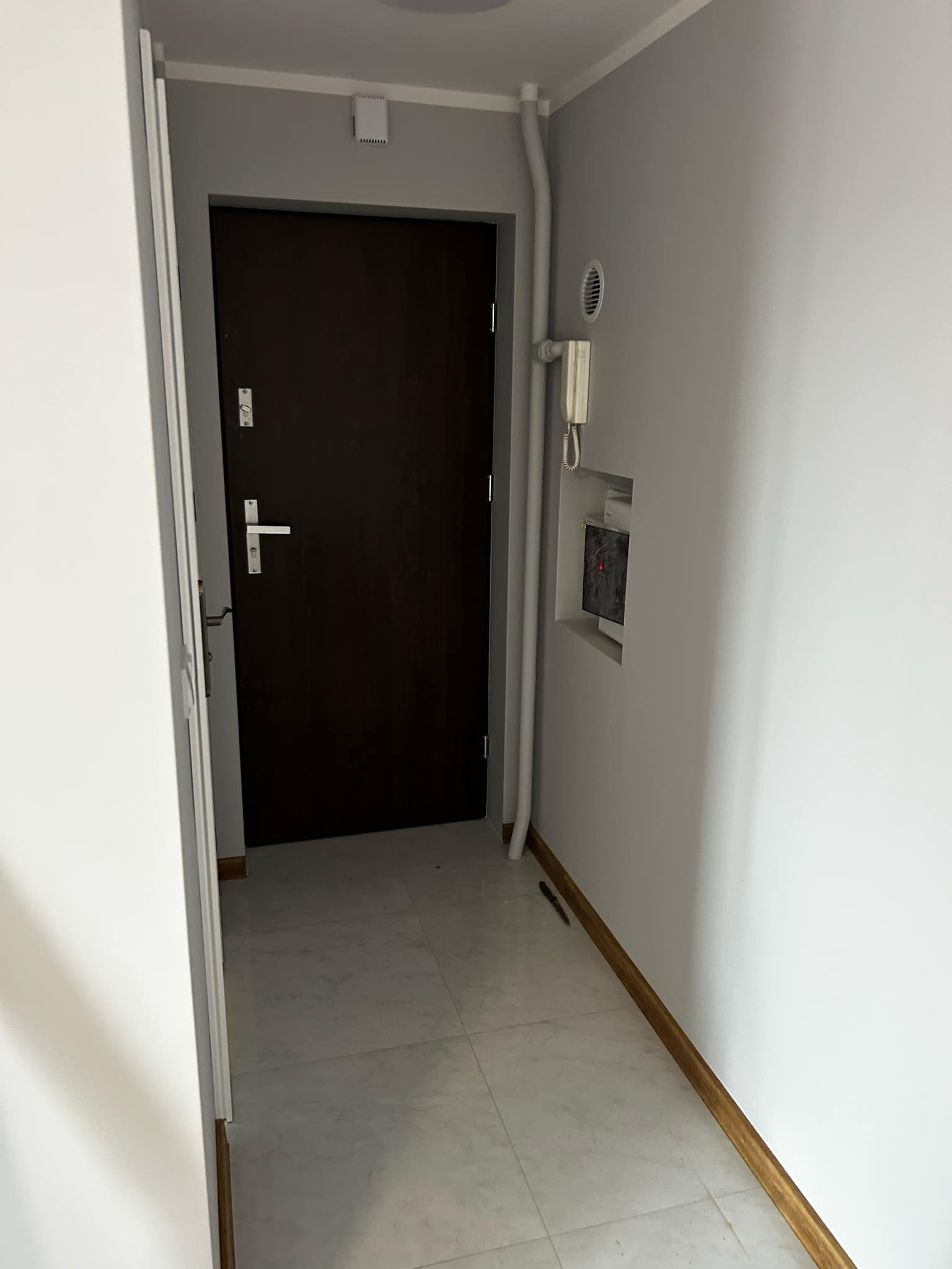 Wykończony korytarz z brązowymi drzwiami i jasną podłogą z płytek. Widoczny domofon i listwy przypodłogowe. Ściany pomalowane na szaro.