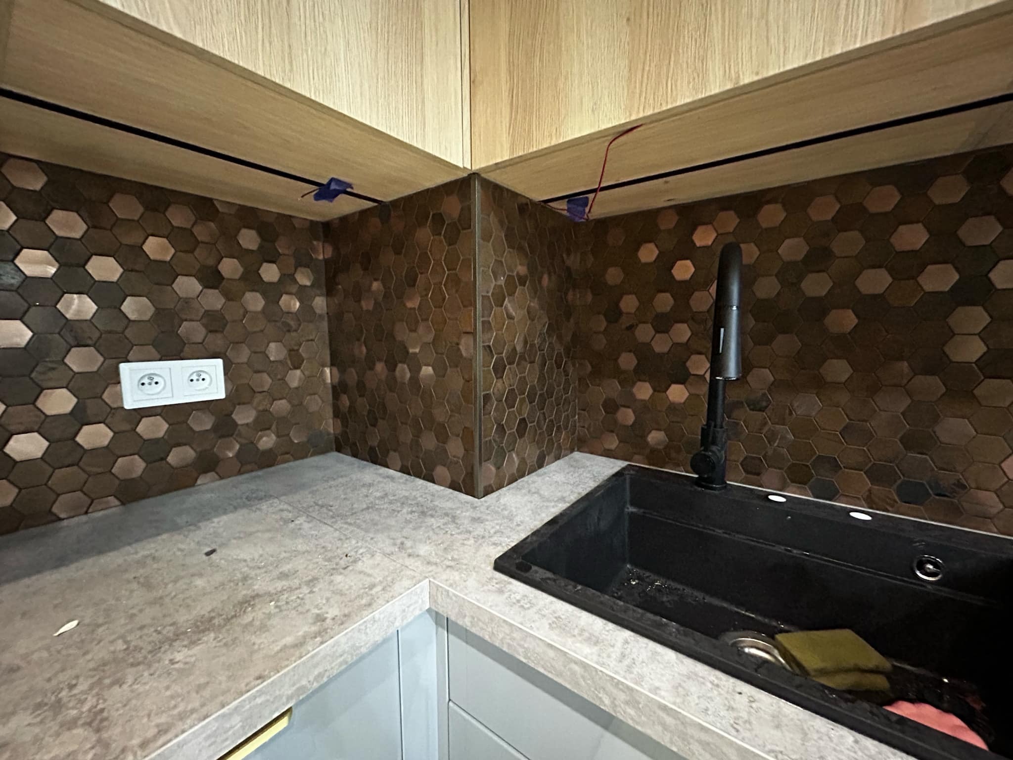 Kuchnia po remoncie: heksagonalne płytki w odcieniach brązu na ścianie nad blatem, czarny zlew i drewniane wykończenie podszafkowe. Widoczne gniazdko elektryczne.