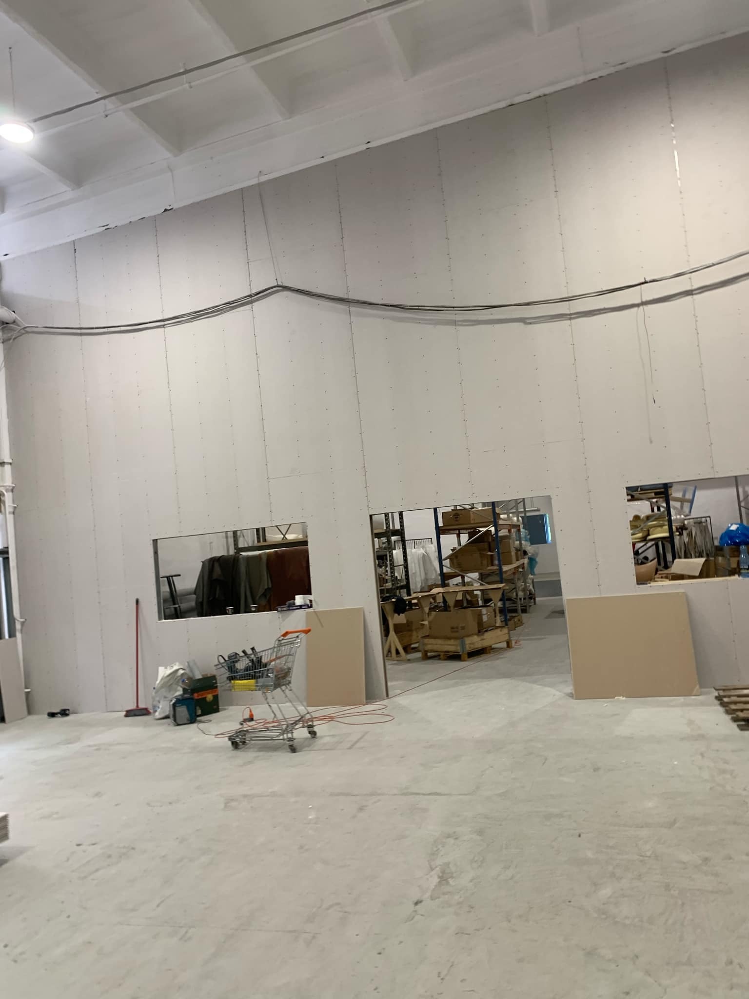 Wnętrze w trakcie wykańczania: ściany z płyt kartonowo-gipsowych z otworami, wózek z narzędziami i materiałami budowlanymi na betonowej podłodze. Widoczne kable pod sufitem.