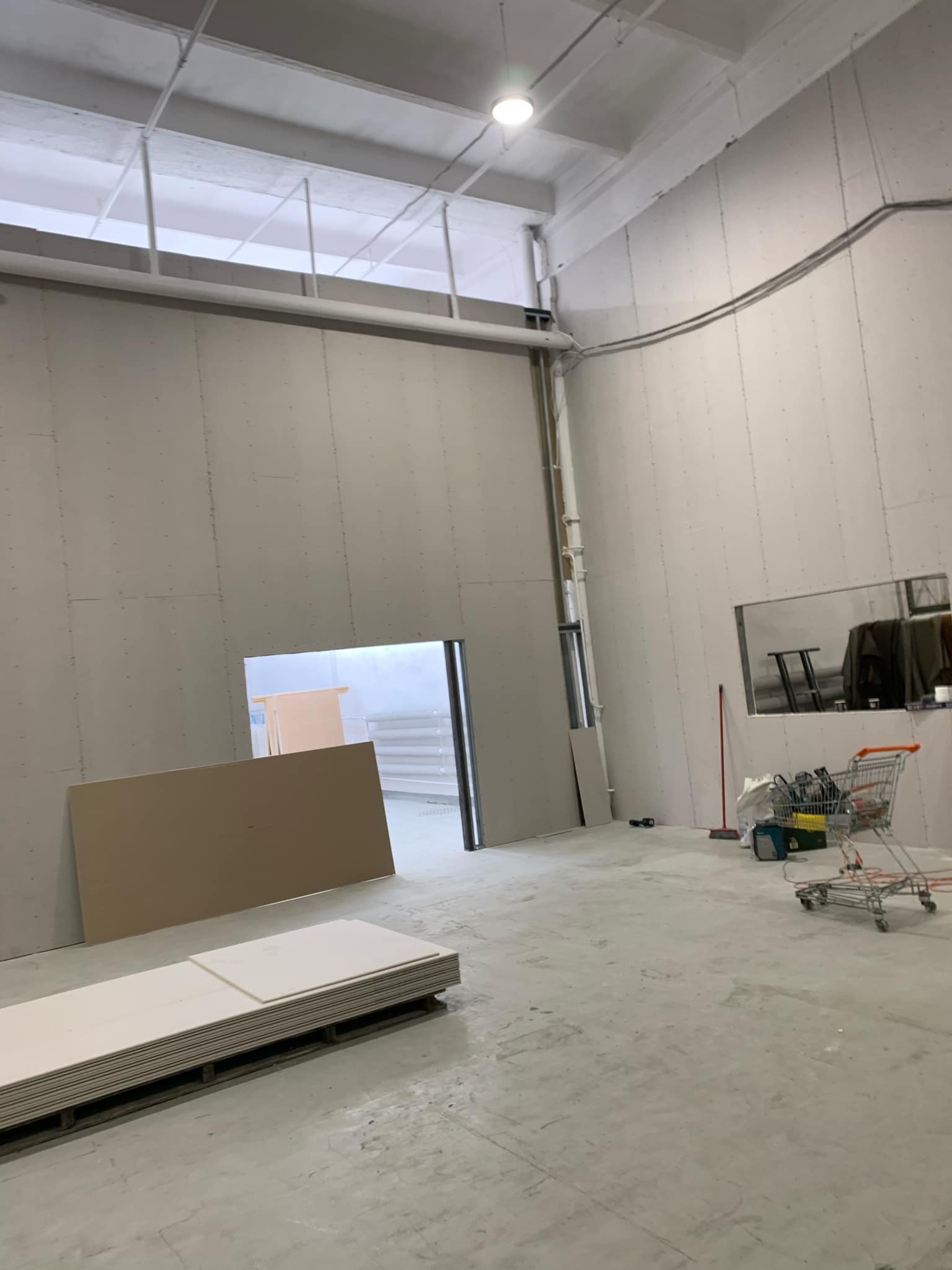 Wnętrze w trakcie wykańczania: ściany z płyt gipsowo-kartonowych, sterta płyt na podłodze, narzędzia i wózek w tle. Widoczne przygotowania do dalszych prac wykończeniowych.